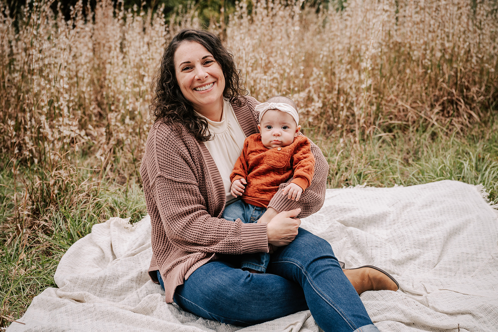 A mom sits on a picnic blanket in a field of tall grass while holding her infant baby in her lap Charlotte Fall Festivals