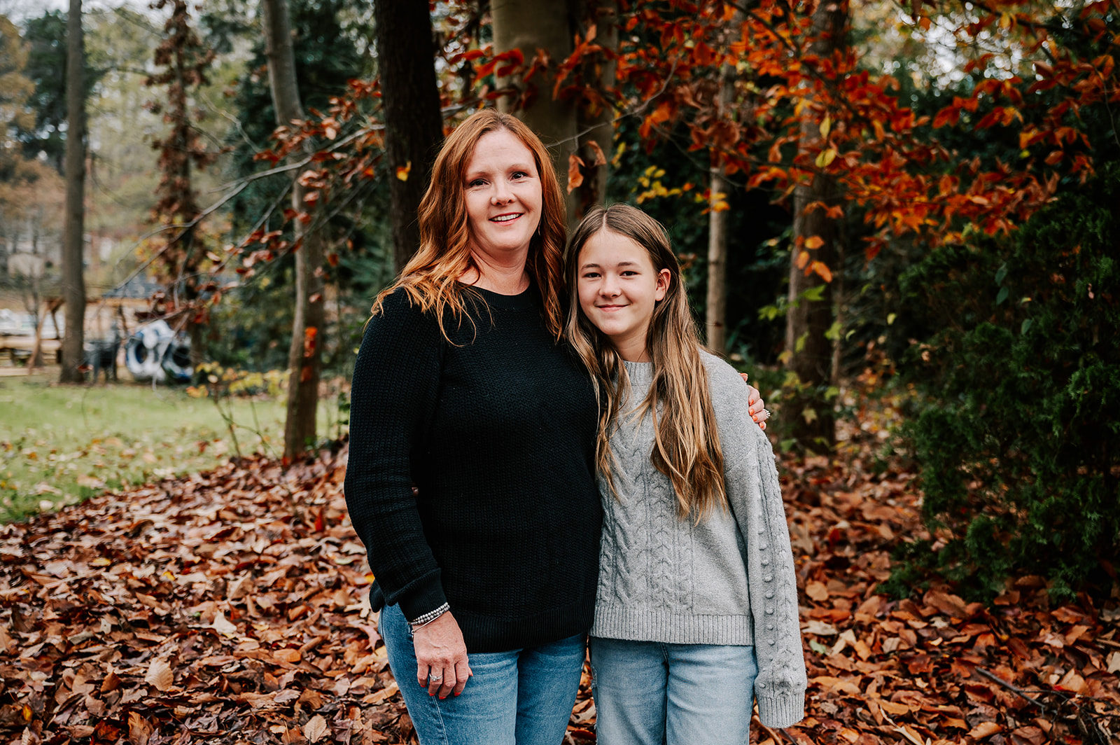 A mom in a black sweater stands by a pile of fallen leaves with her arm around her teen daughter in a grey sweater