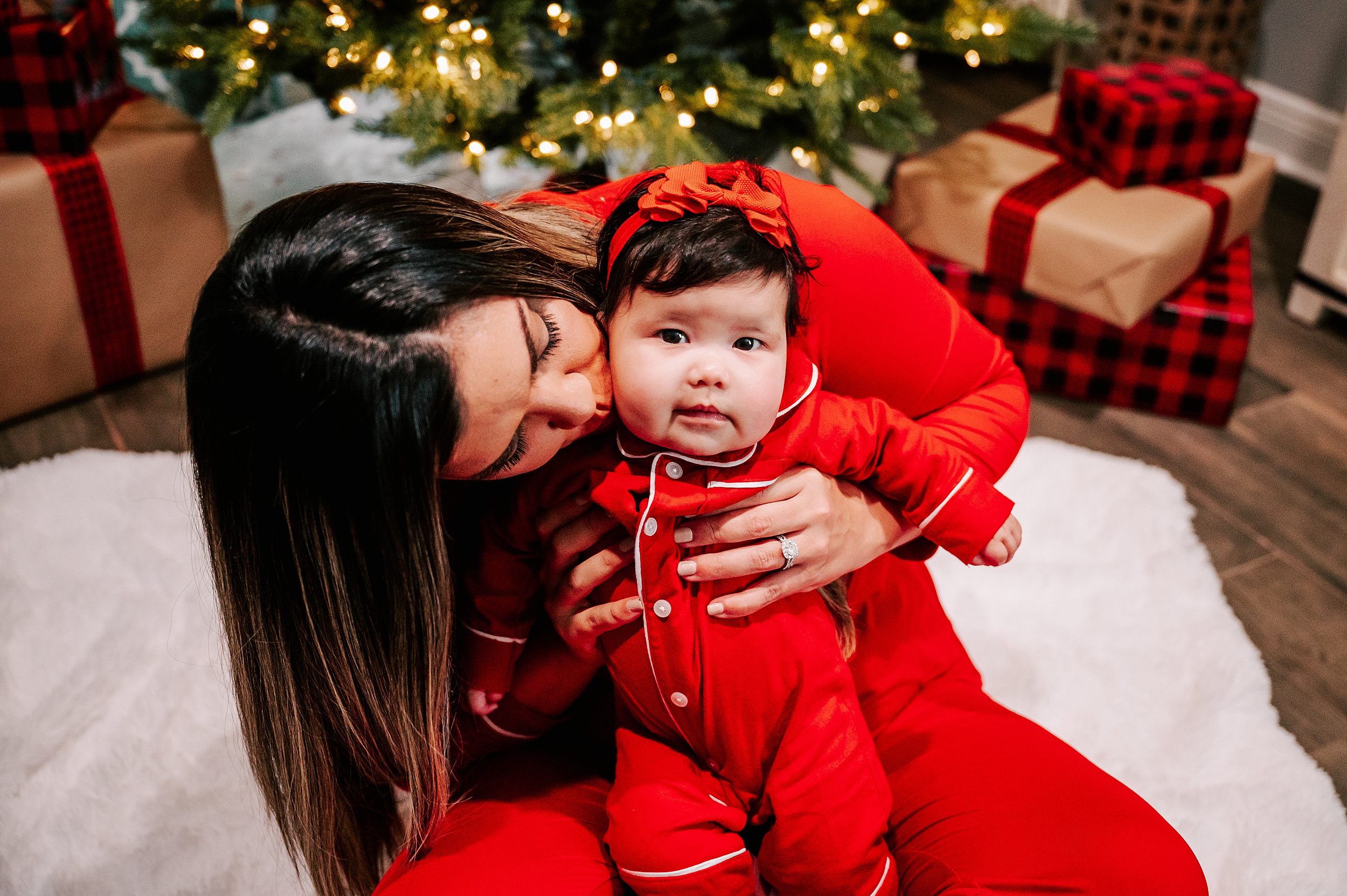 A mom kisses the cheek of her infant daughter while sitting in front of their christmas tree