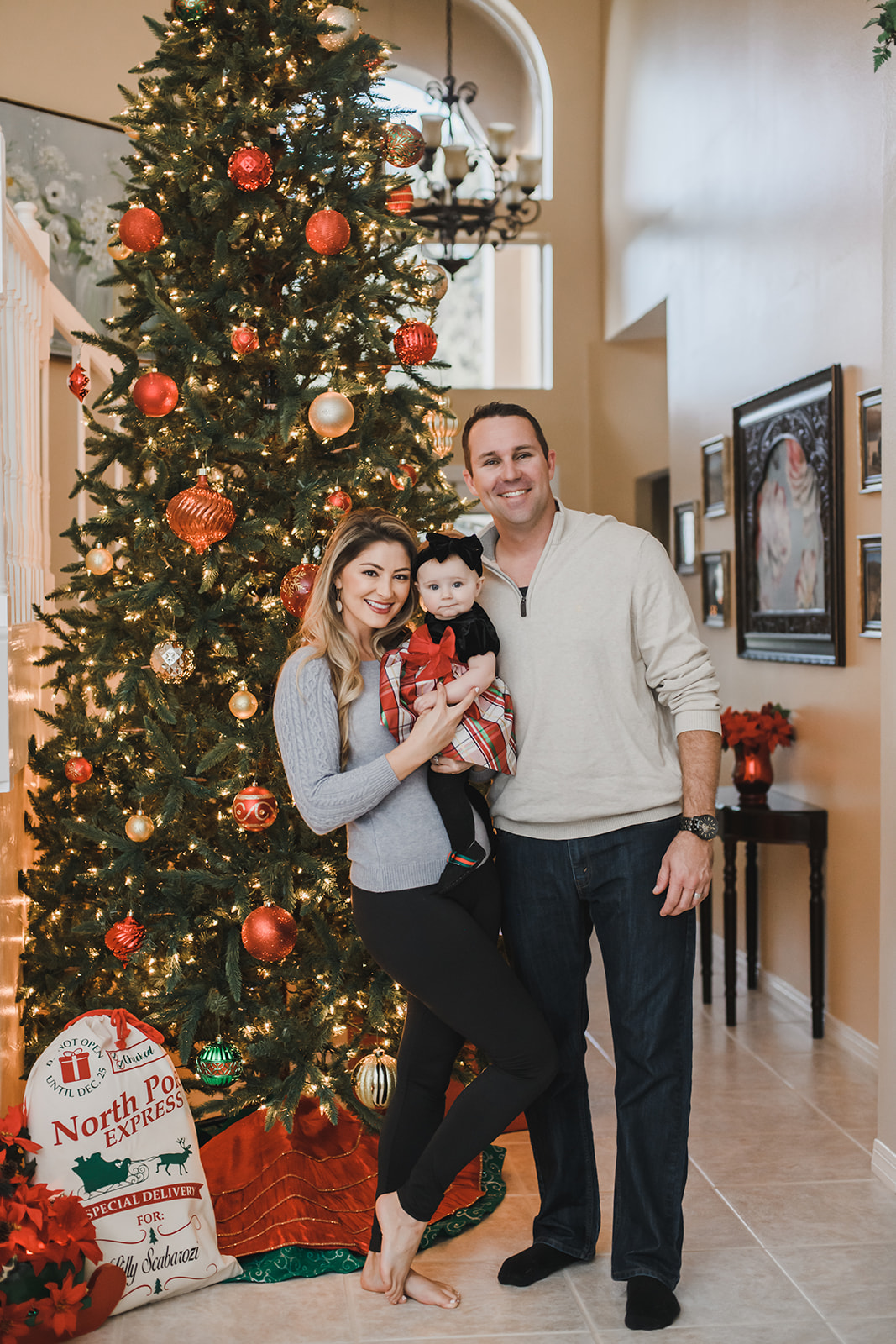 A mom and dad stand together by their christmas tree while holding their infant daughter