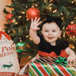 An infant girl sits in a black dress opening presents under the tree Christmas Tree Farms Near Winston-Salem