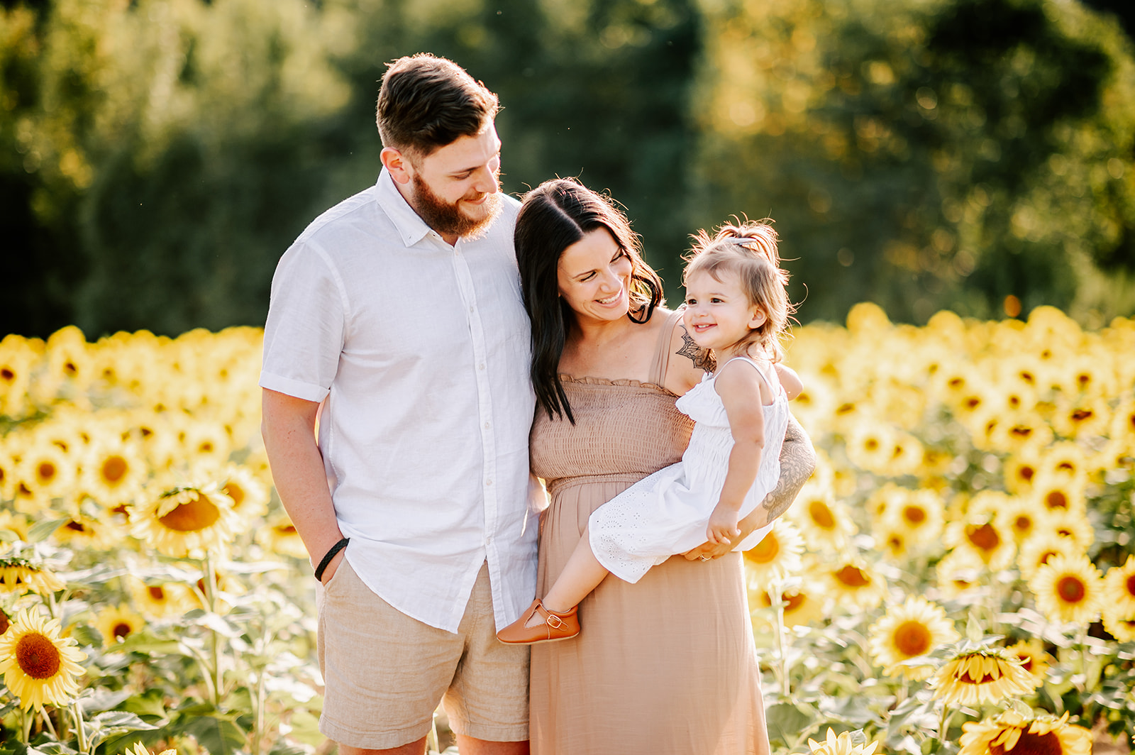 A mom and dad smile down at their smiling daughter while standing in a field of sunflowers