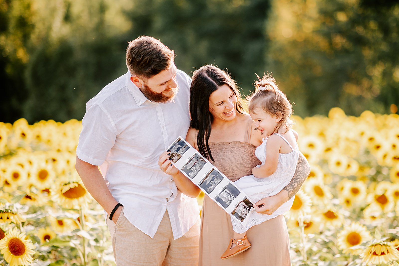 A pregnant mother holds her daughter on her hip while showing her sonogram images with dad while standing in a sunflower farm