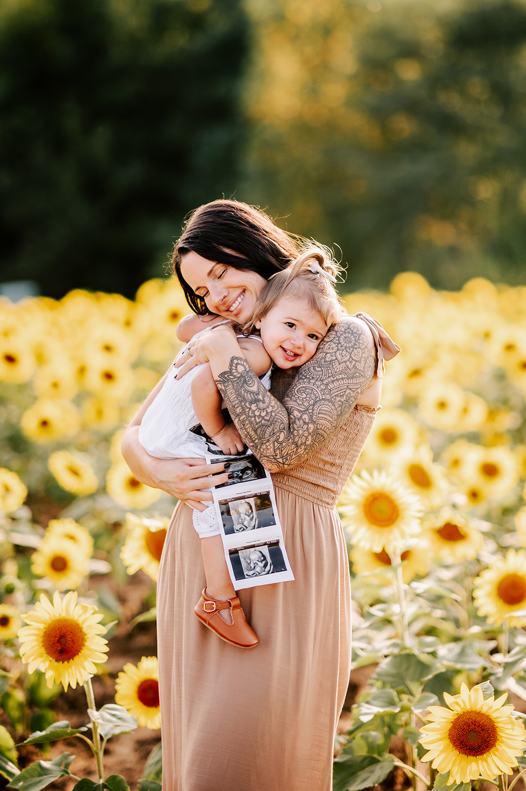 A pregnant mom holding her sonograms hugs her smiling toddler daughter while standing in a field of sunflowers at Dogwood Farms