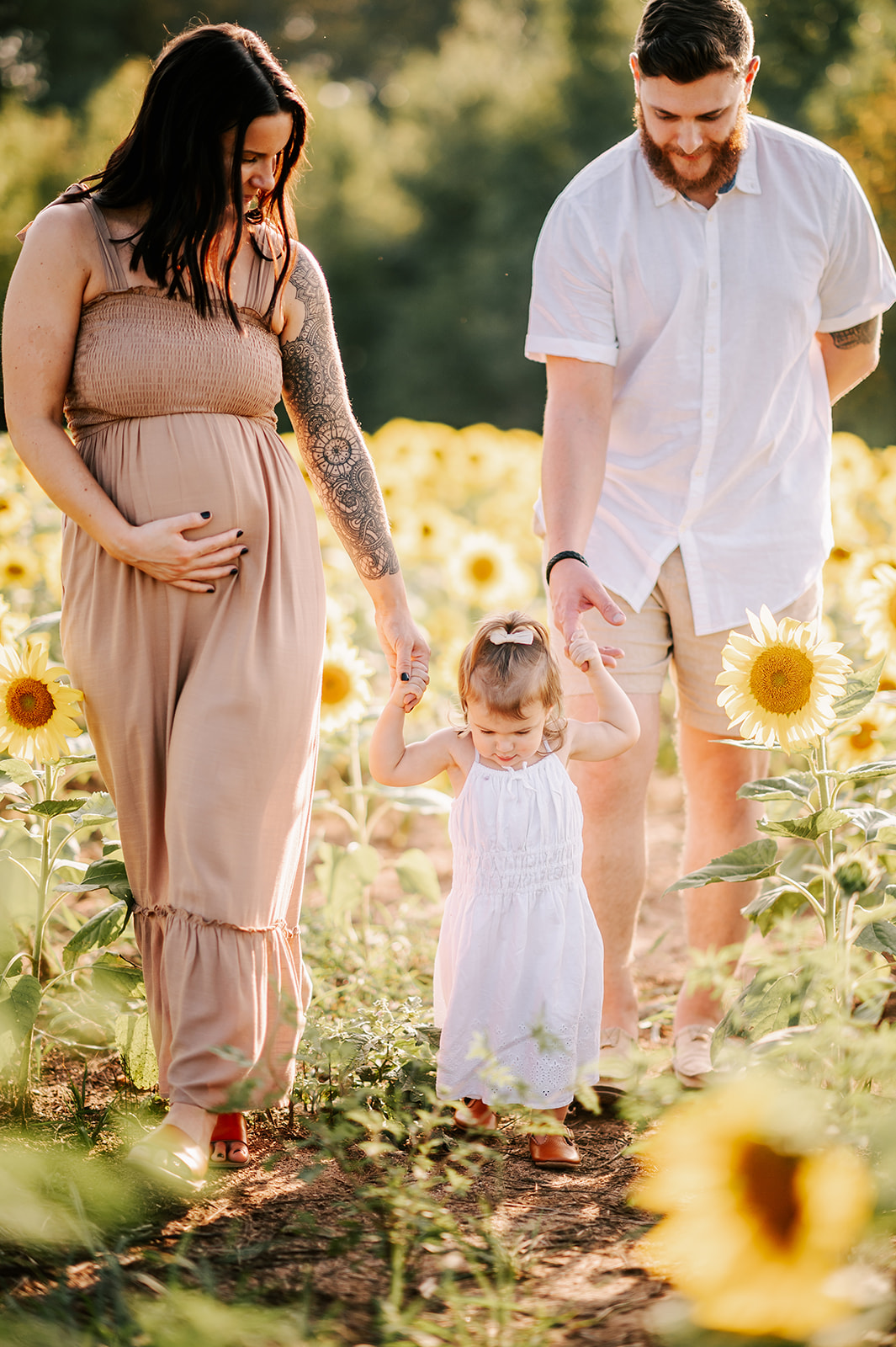 A pregnant mom and dad lead their toddler daughter in a white dress through a field of sunflowers at Dogwood Farms