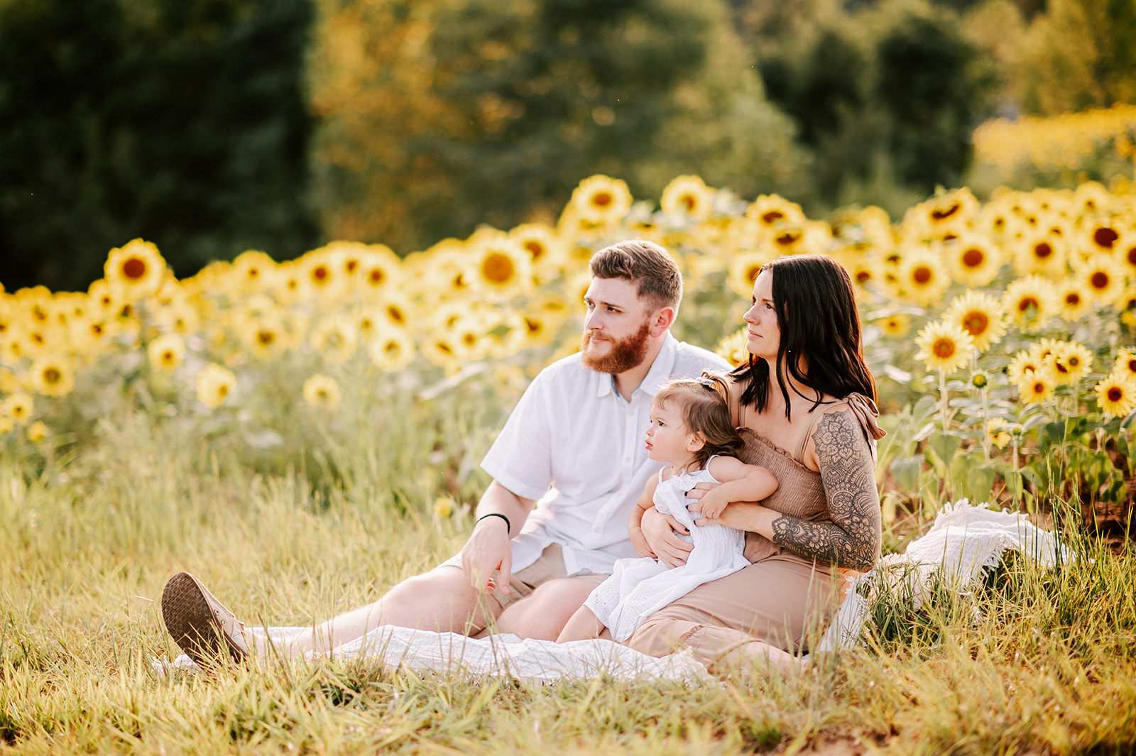 A pregnant mother sits on a picnic blanket with her toddler daughter in her lap and husband at her side in a field of sunflowers