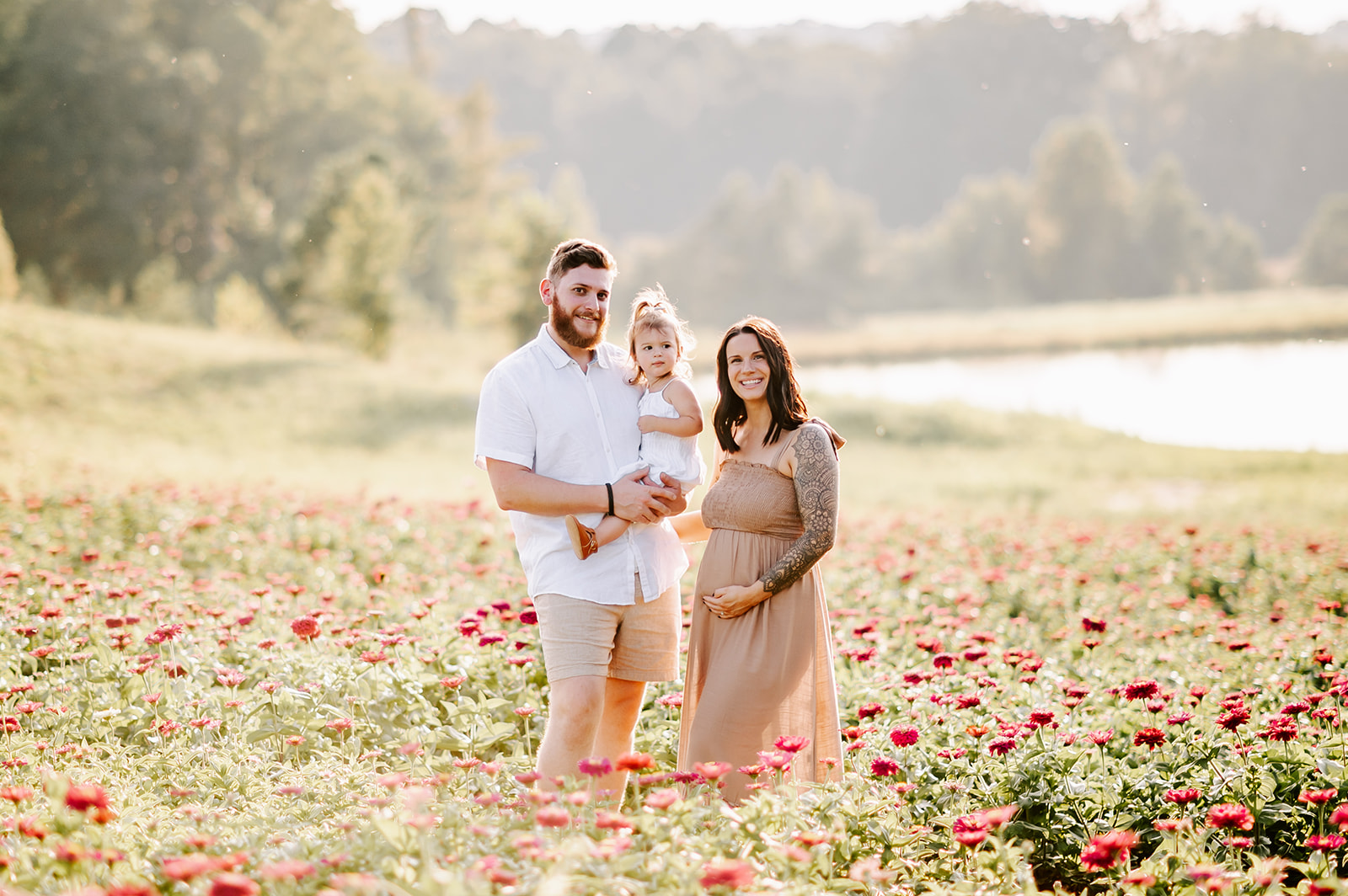 A pregnant mother stands in a field of flowers on a farm with her husband holding their toddler daughter on her hip