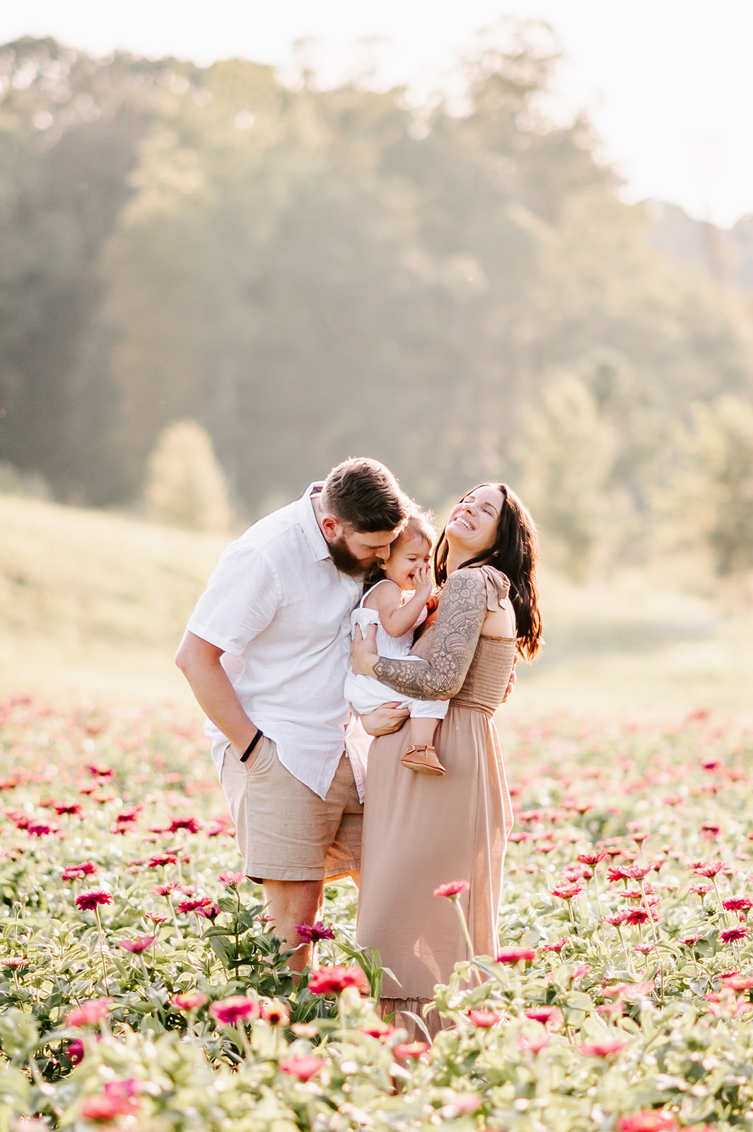 A toddler girl in a white dress sits on her pregnant mom's bump while dad leans in to play with them while standing in a field of flowers