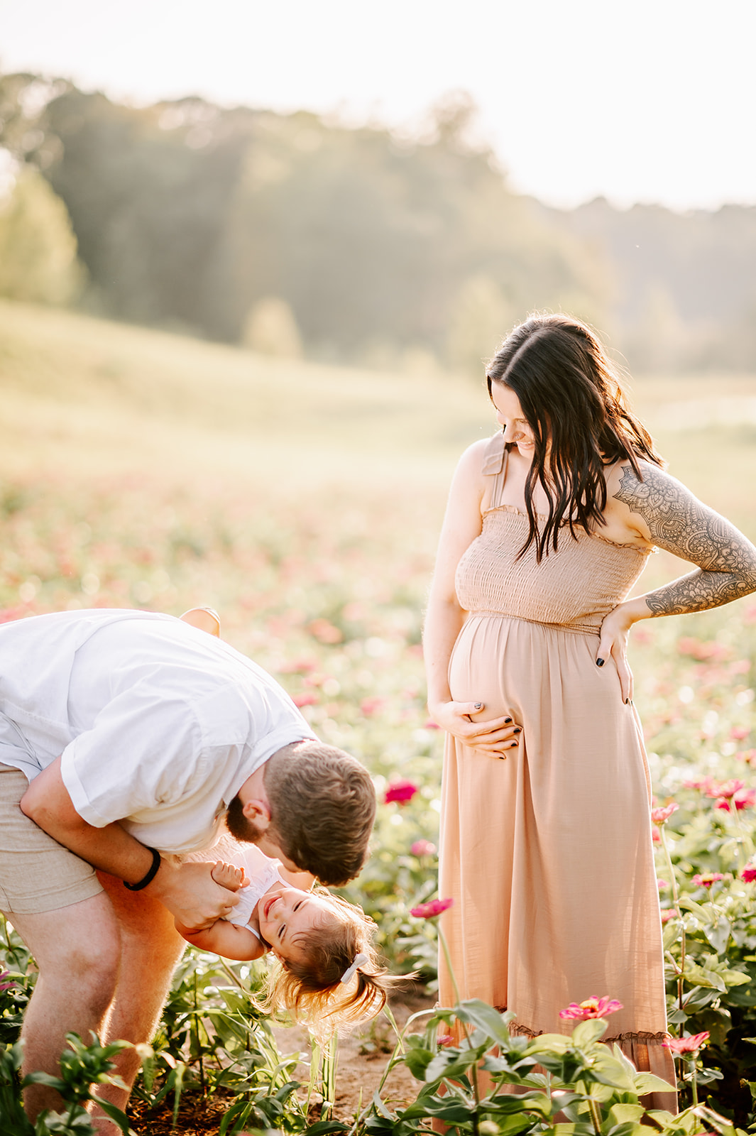 A pregnant mother holds her bump while smiling down at her husband flipping and playing with their toddler daughter in a field of flowers at Dogwood Farms