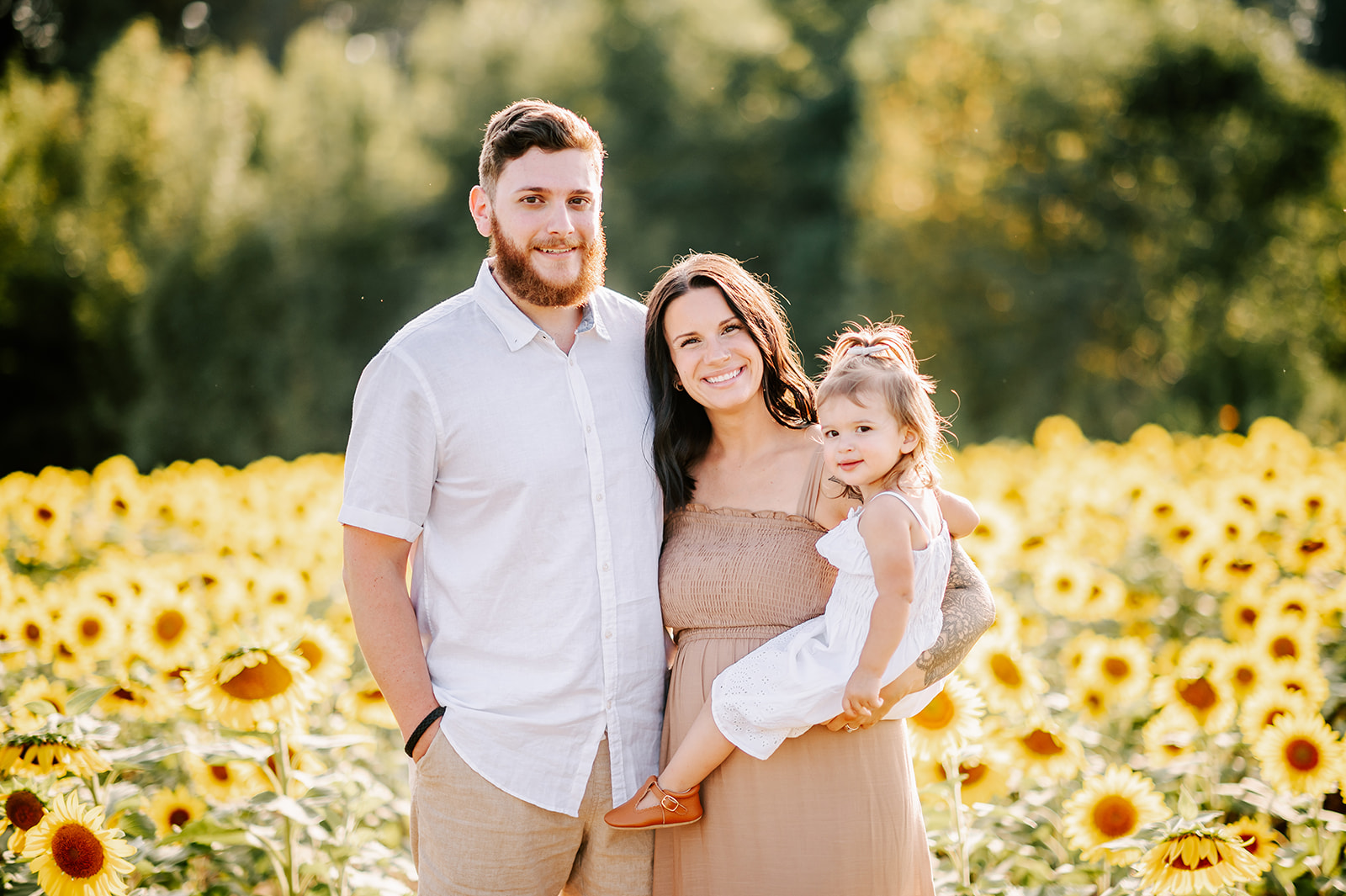 A mother and father stand in a field of sunflowers at Dogwood Farms while their toddler daughter sits on mom's hip