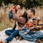 A mom and dad play and lift their infant twin daughters while sitting on a blanket in some tall golden grass Greensboro Apple Picking