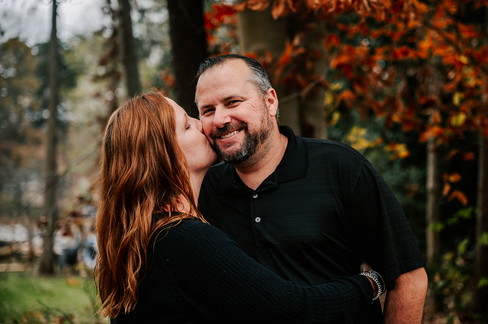 A mom hugs and kisses the cheek of her husband while standing in a park in fall Raleigh Fall Activities