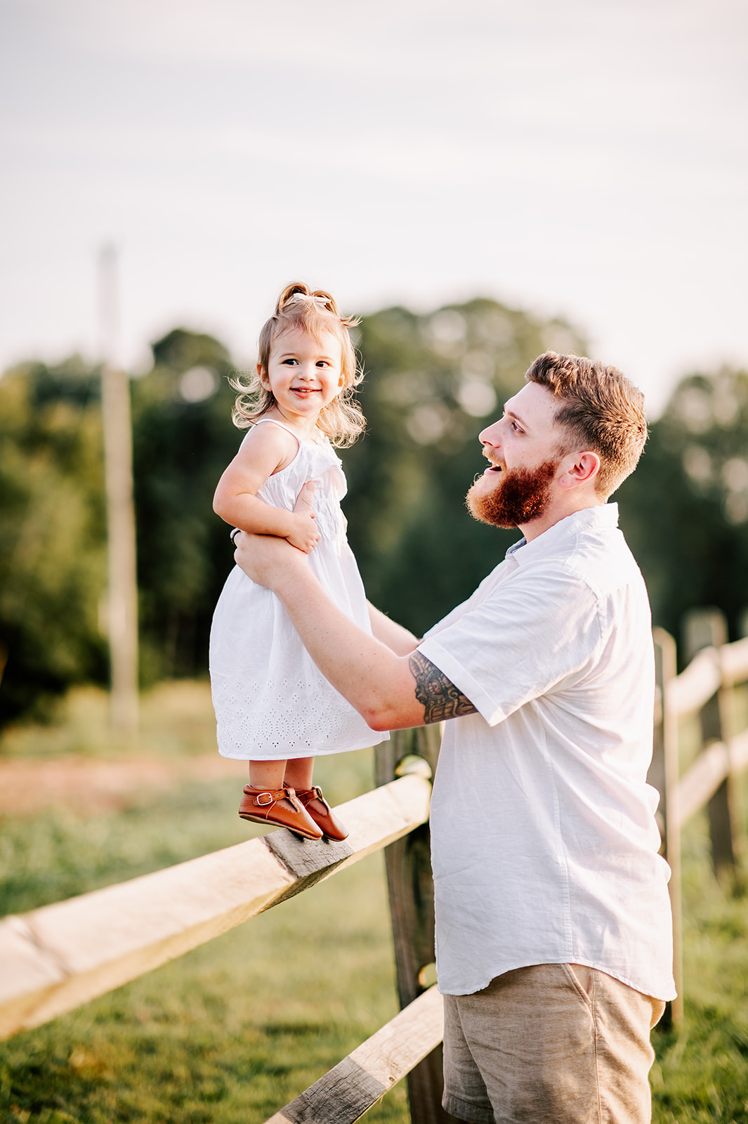 A toddler girl in a white dress stands on a wooden fence while dad holds her