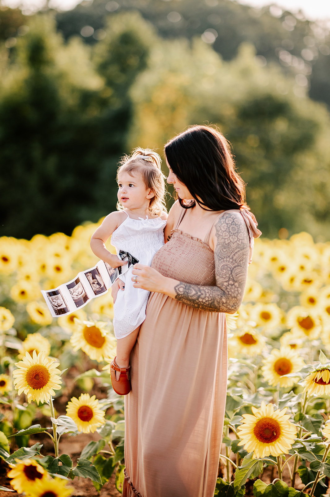 A mom in a beige maternity gown stands in a field of sunflowers with her toddler on her hip in a white dress holding a string of sonograms during Asheville Fall Activities