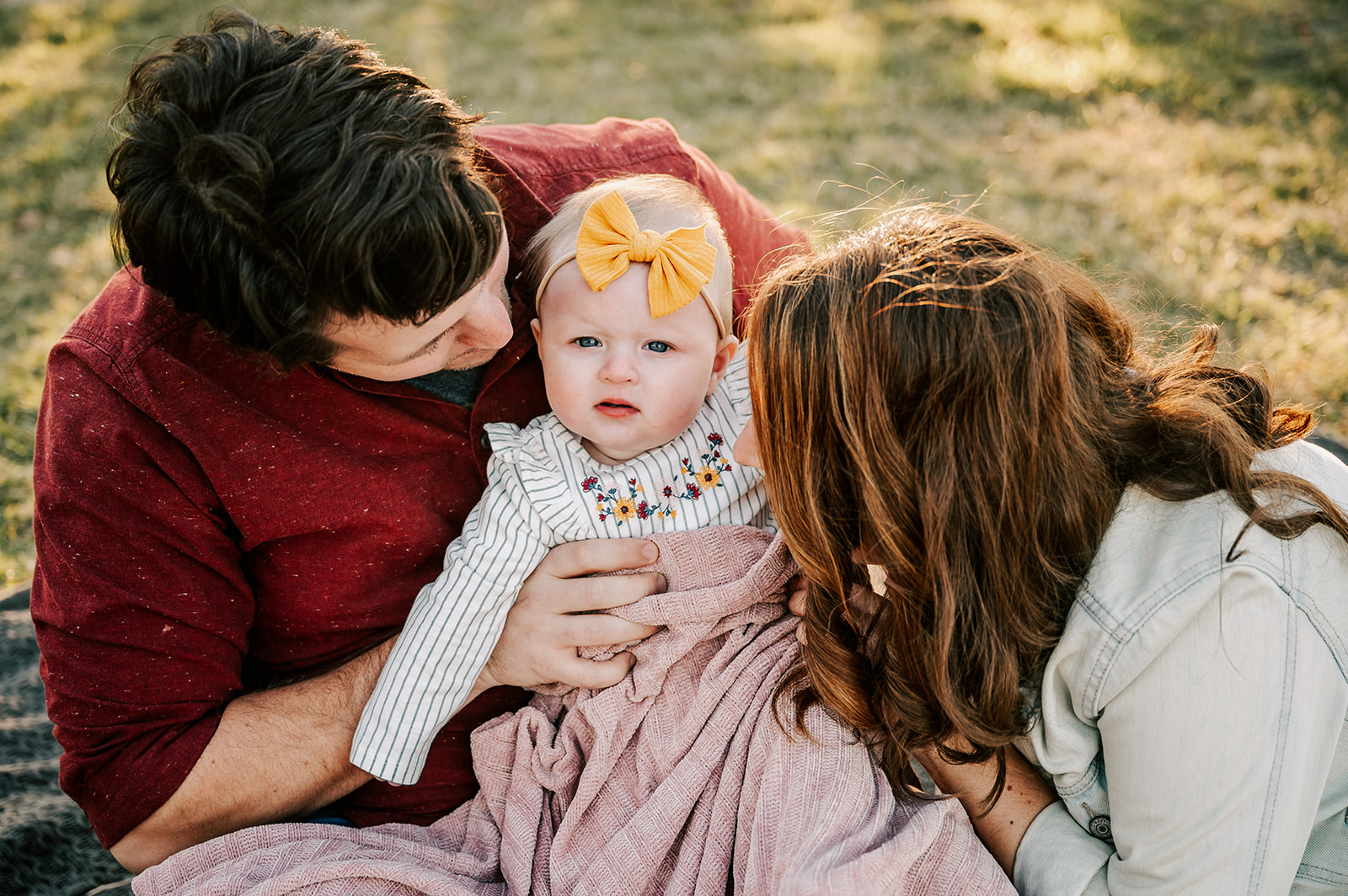 A mom and dad smile down at and tickle their infant daughter in dad's lap in a park while sitting on a blanket