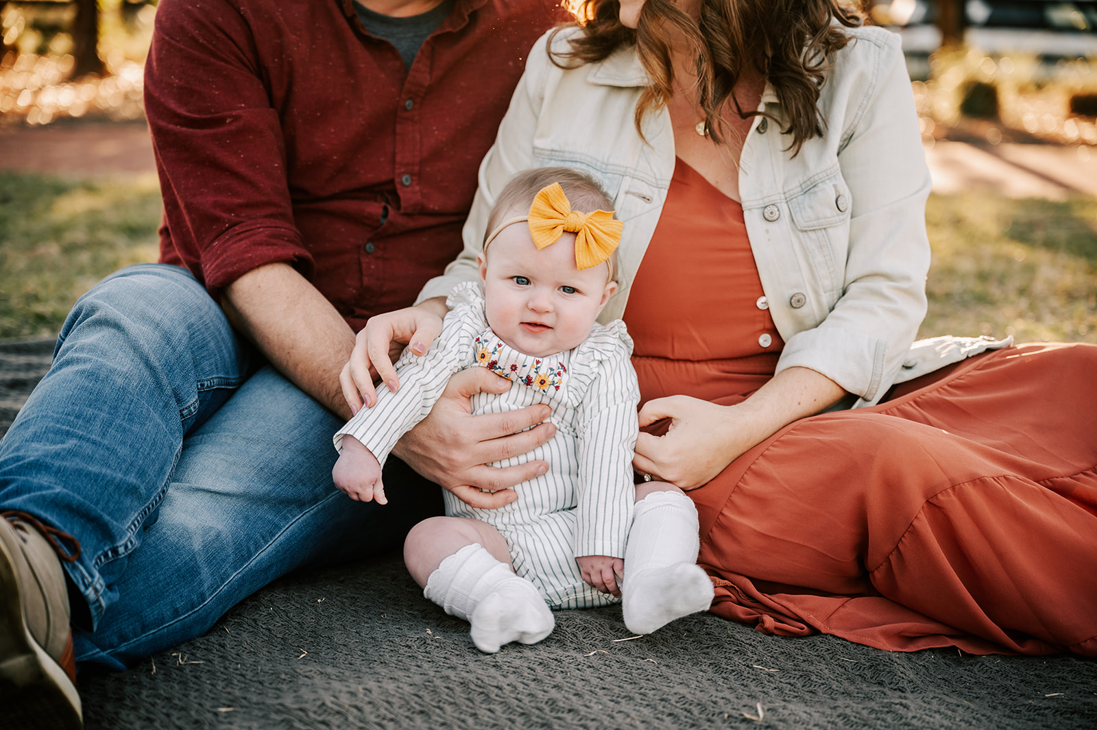 An infant girl in a white striped onesie and yellow boy sits on a blanket with mom and dad in a park at sunset