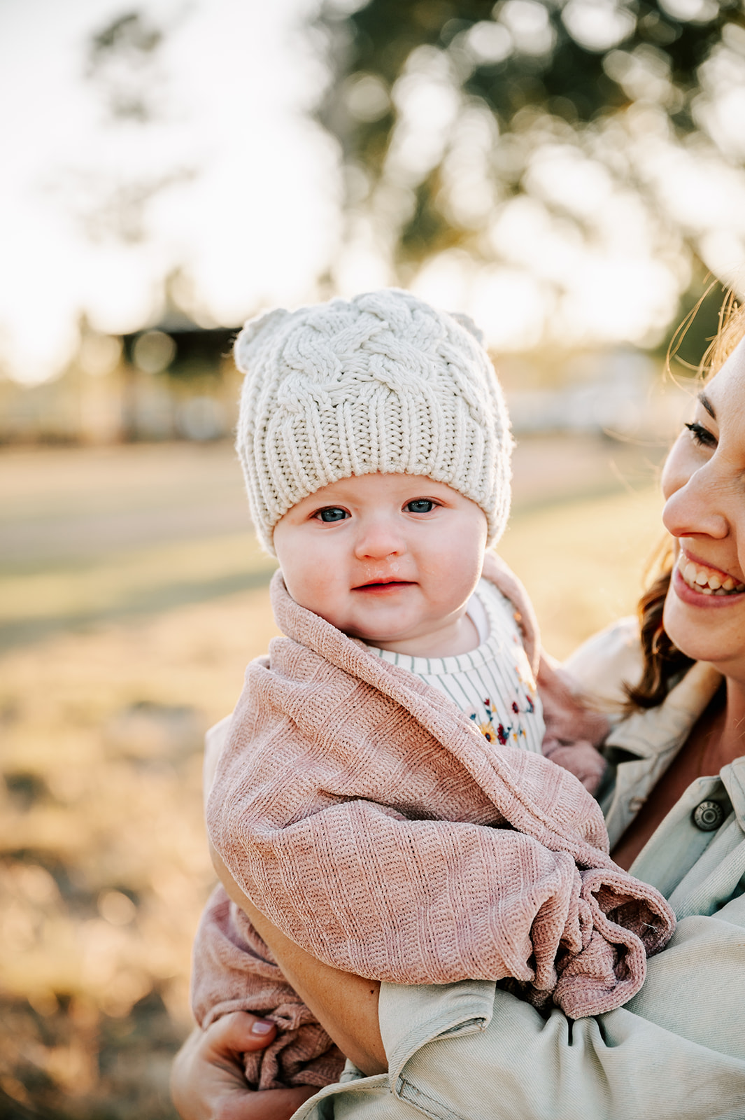 An infant in mom's arms smiles in a white beanie and wrapped in a pink blanket during some Charlotte Fall Activities