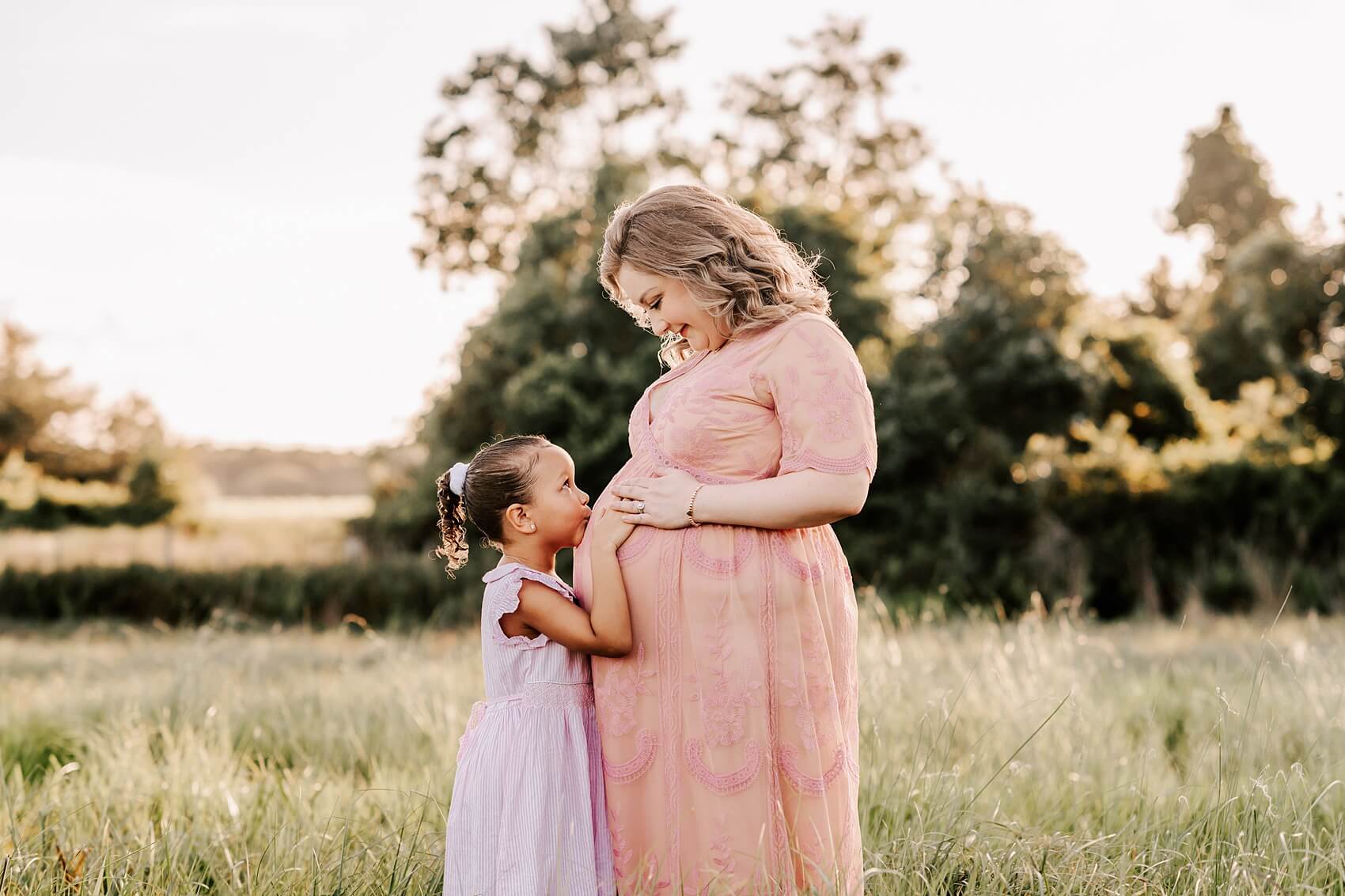 A mom to be in a pink lace maternity gown stands in a field as her daughter kisses her belly in a purple dress growing years learning center