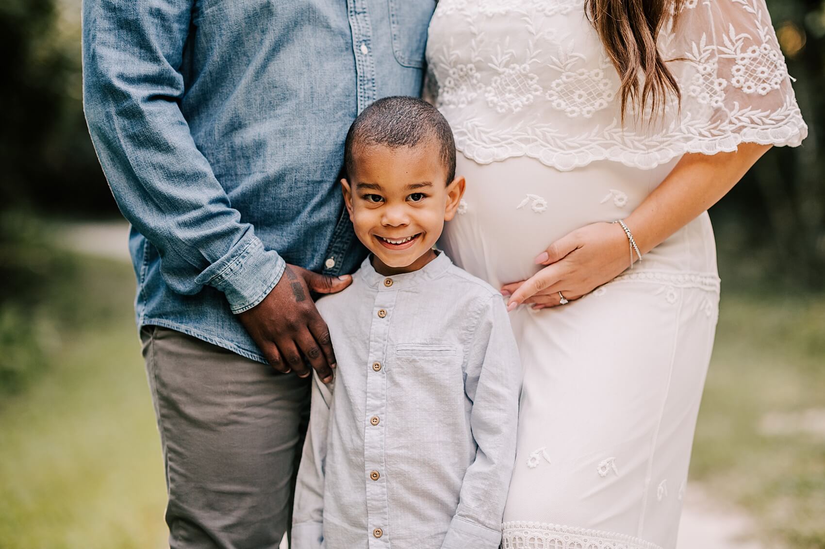 A young boy smiles big in a grey shirt between his pregnant mom in a white lace maternity gown and dad in a denim shirt and grey pants in a park after attending growing years learning center
