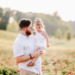 A father in a white shirt stands in a field of flowers holding his curious toddler daughter in a white dress on his hip before doing some North Carolina Apple Picking
