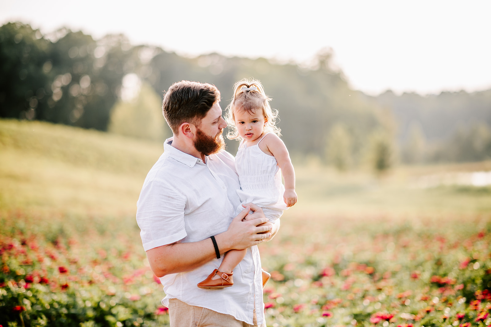 A father in a white shirt stands in a field of flowers holding his curious toddler daughter in a white dress on his hip before doing some North Carolina Apple Picking