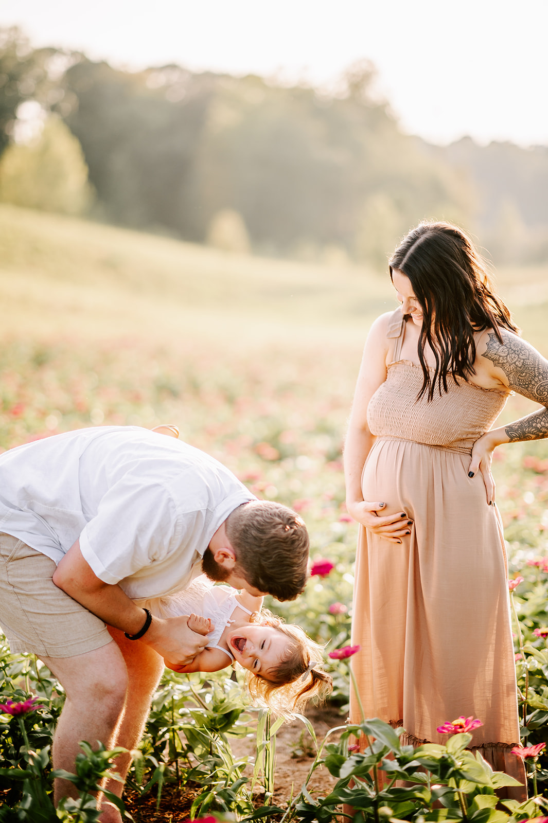 A father plays and dips his toddler daughter in a field of flowers with pregnant momma smiling on in a beige maternity dress before doing some North Carolina Apple Picking