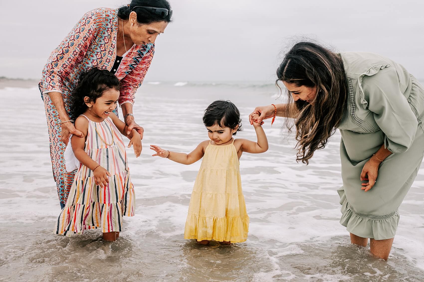 A mother and grandma walk through the shallow water on a beach while playing with two toddler girls in dresses tots to teens raleigh