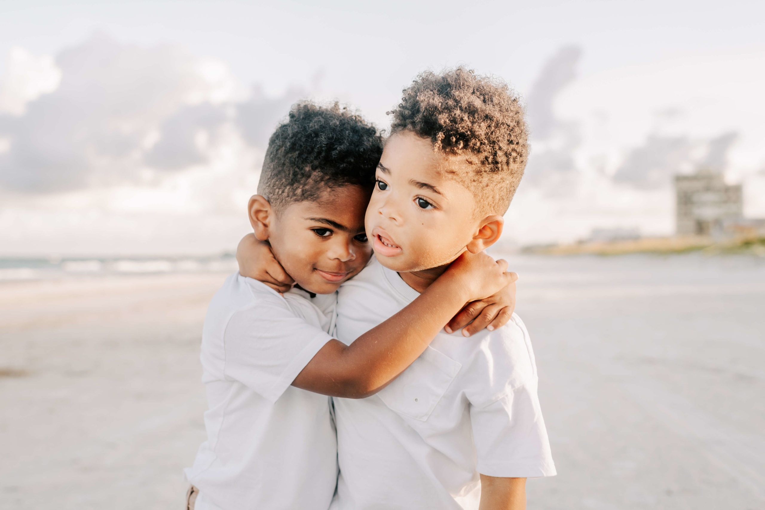 Toddler twin boys hug each other while wearing white shirts on a beach at sunset toys and co