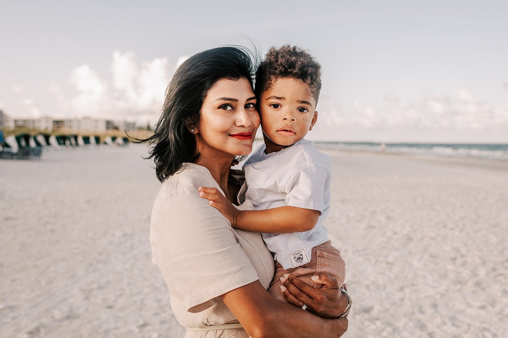 A mother in a beige dress stands on a beach holding her toddler son in a white shirt and red pants