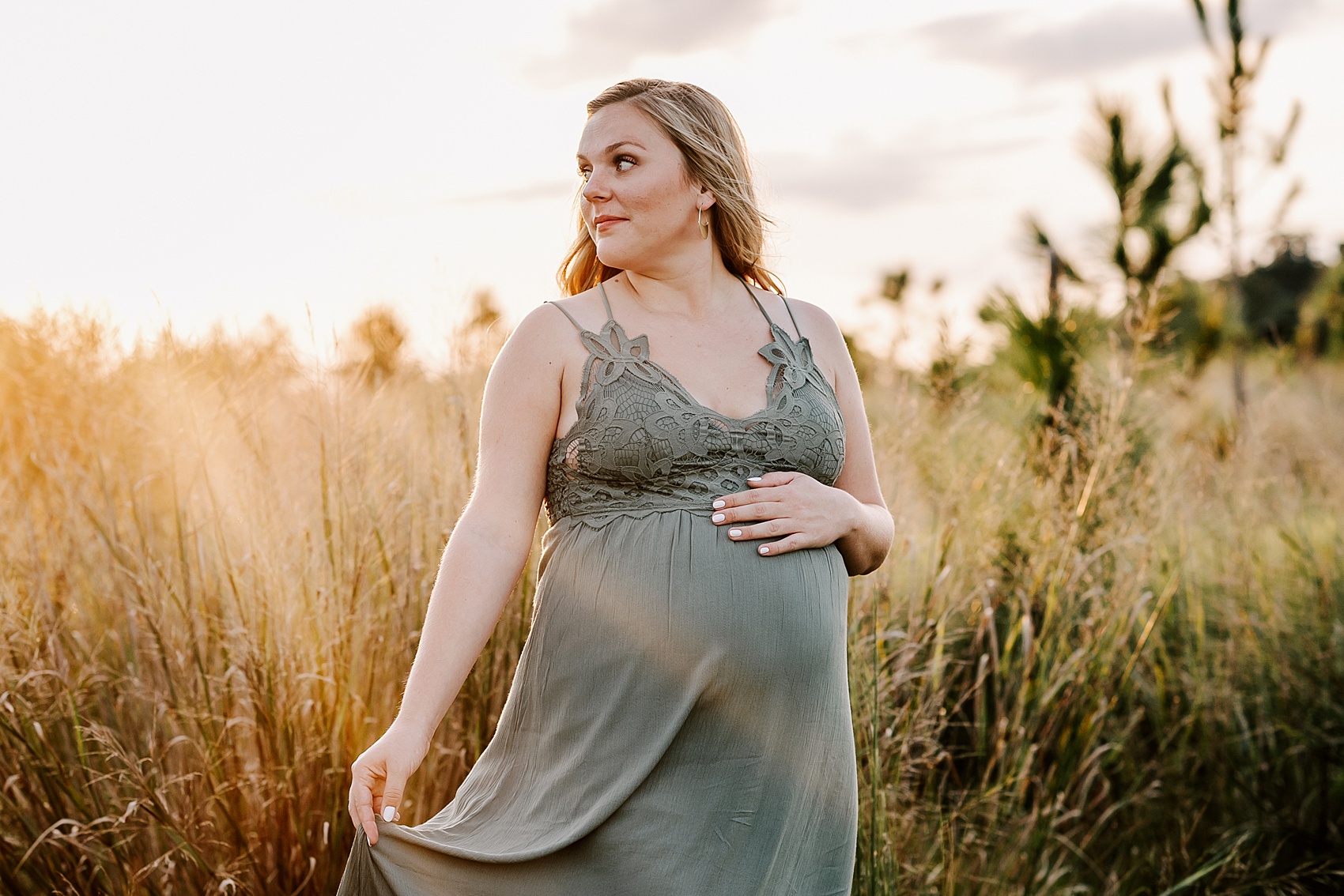 A mom to be in a green maternity dress plays with her dress as she walks through a field of tall grass at sunset