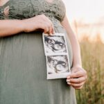 Details of a sonogram being held by a mom to be in a sage green dress standing in a field of tall grass in front of her bump wake forest obgyn shepard street