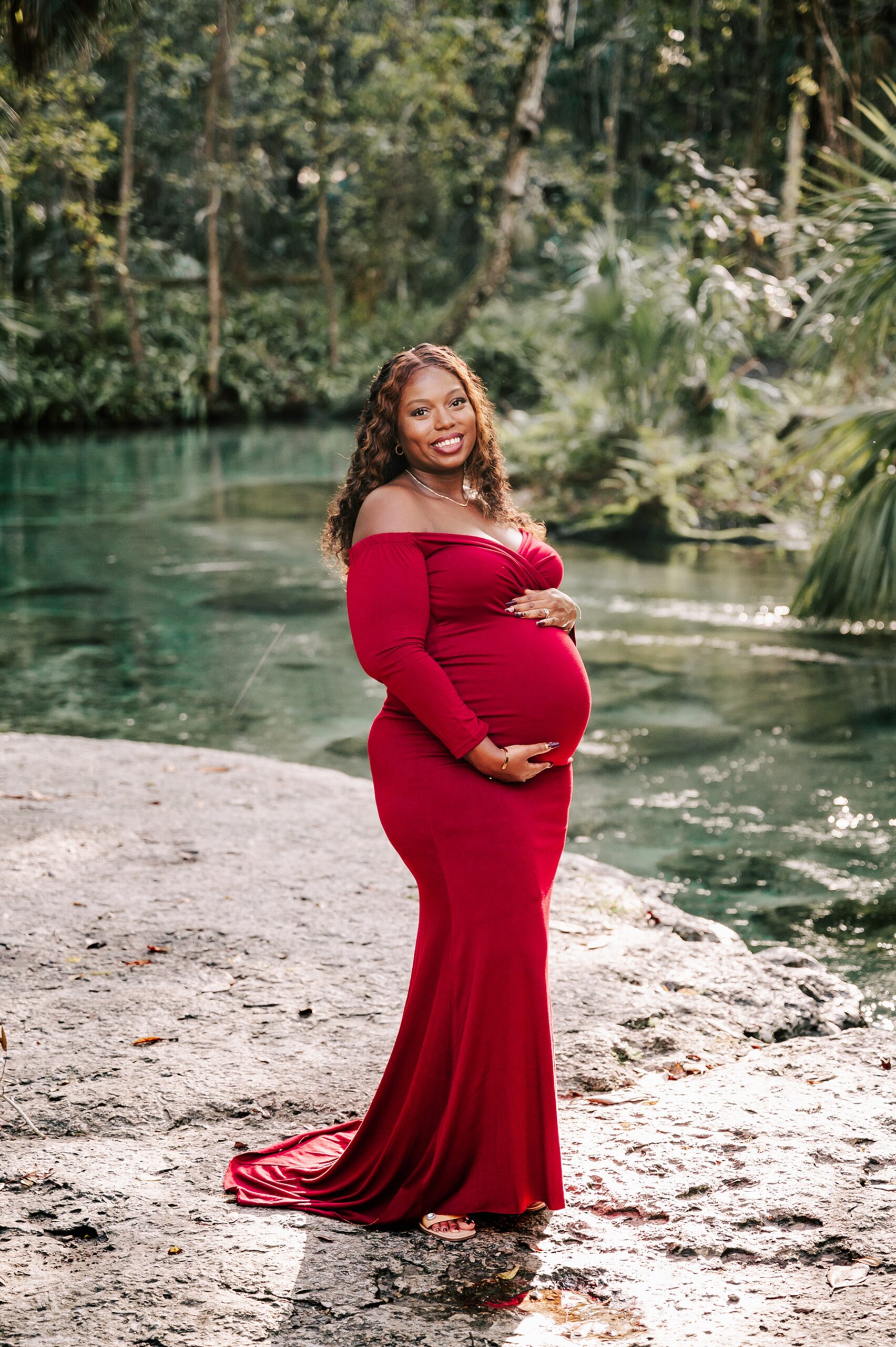 A mother to be stands on the edge of a river holding her bump in a red maternity gown after visiting Elements of Bodywork