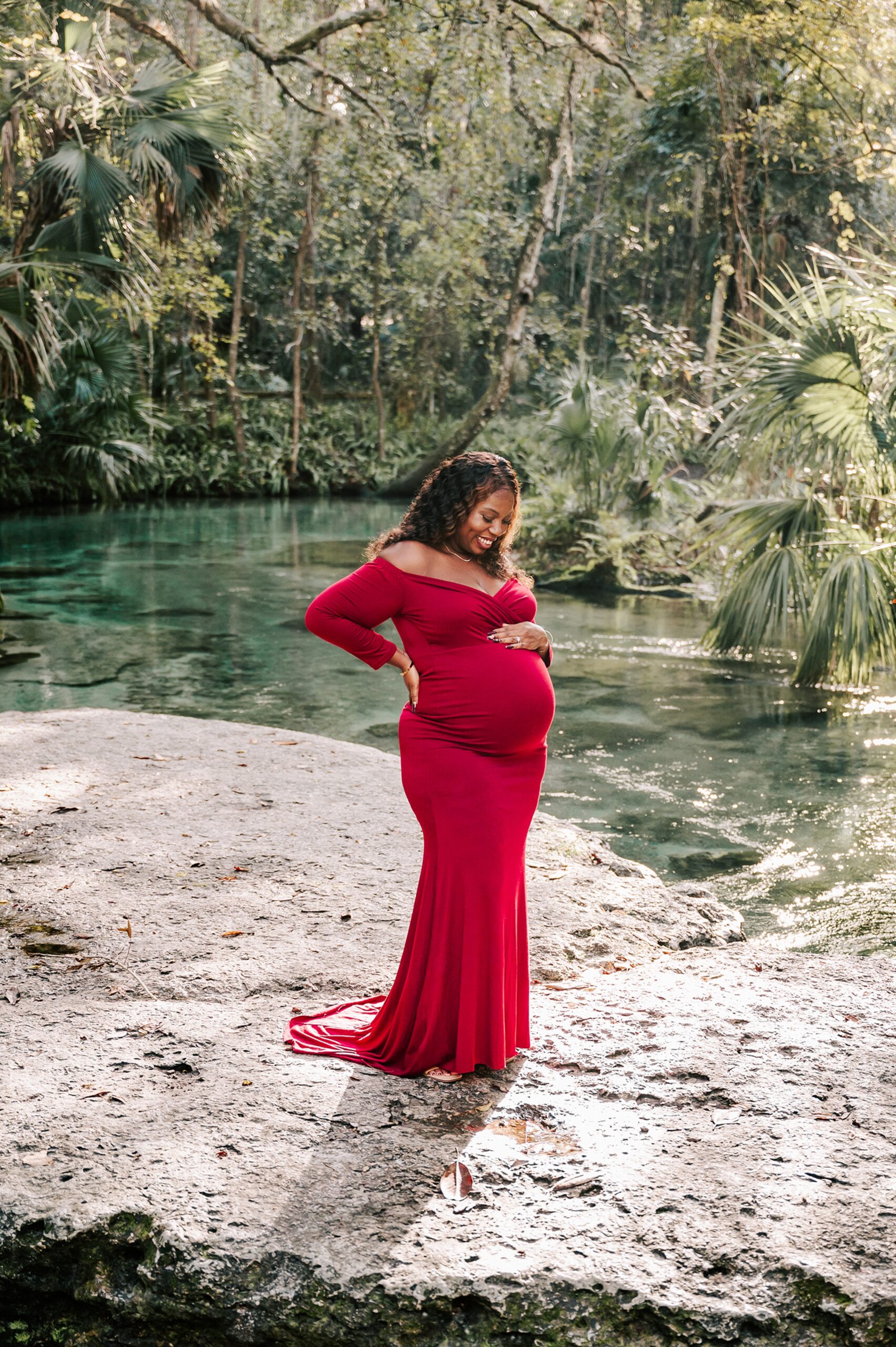 A mother to be stands on a rock in a river smiling down at her bump in a red maternity gown after visiting Elements of Bodywork