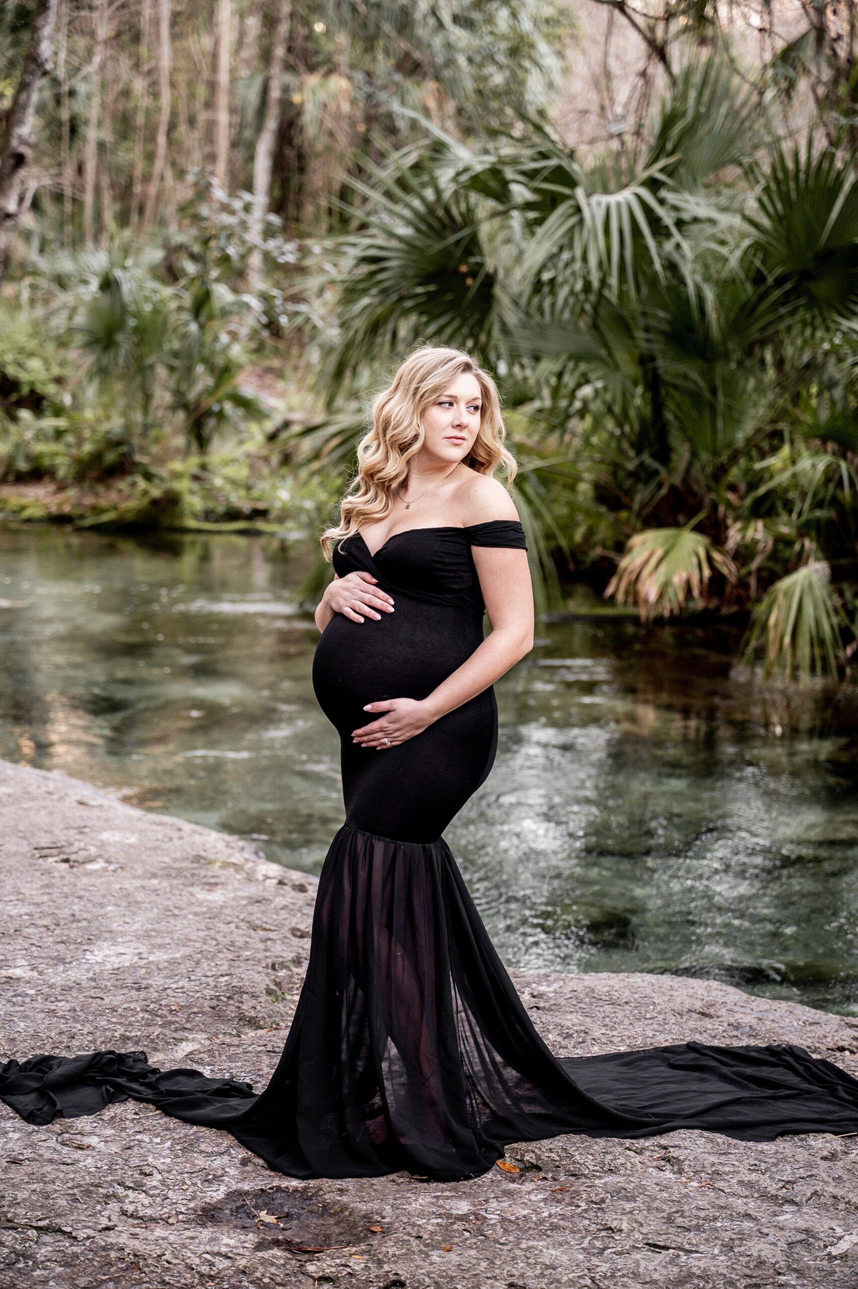 A mother to be glances over her shoulder while standing by a river in a black maternity gown