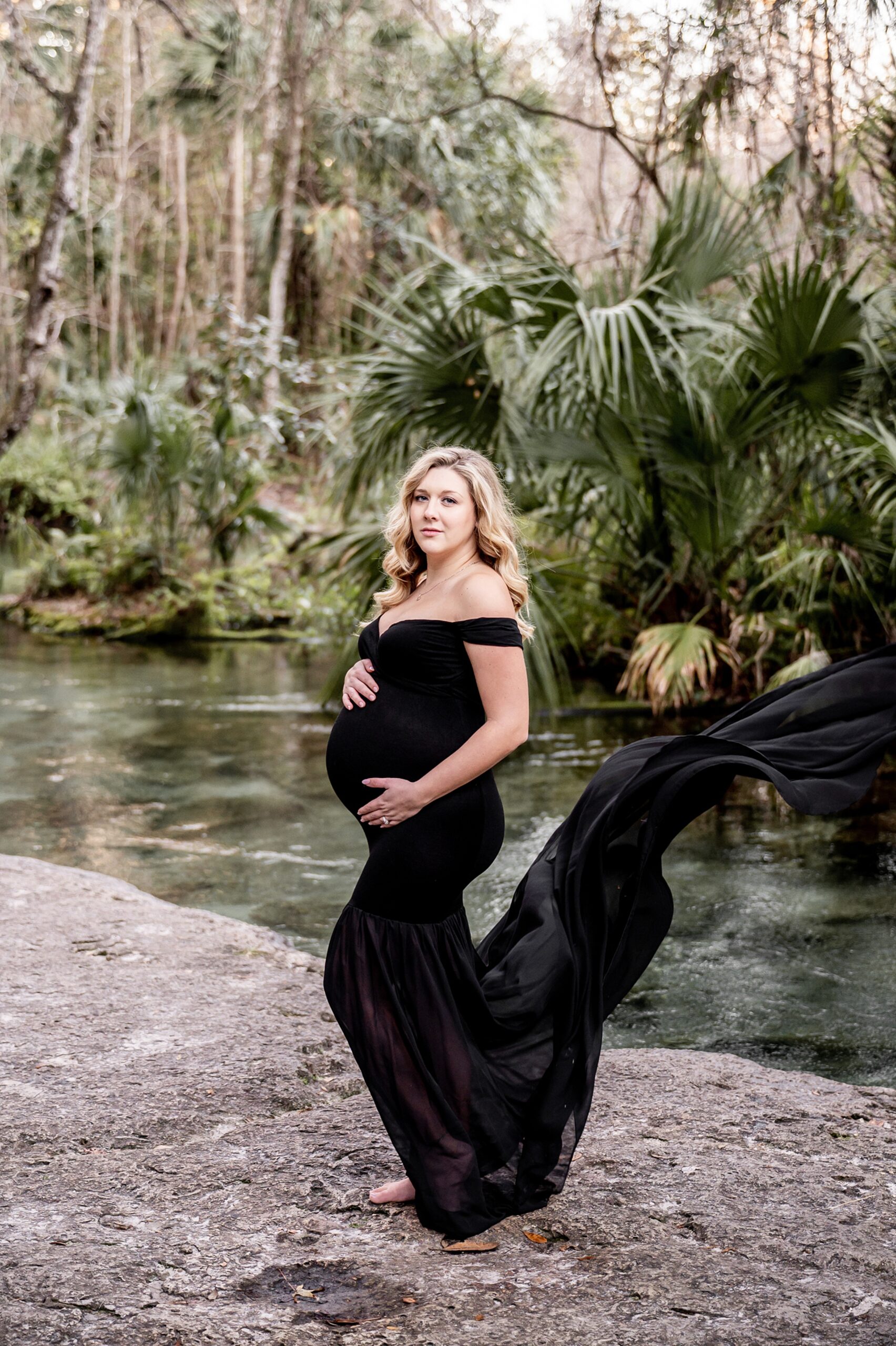 A mother to be in a long flowing black maternity gown stands with hands on the bump on the edge of a river after using duke prenatal care