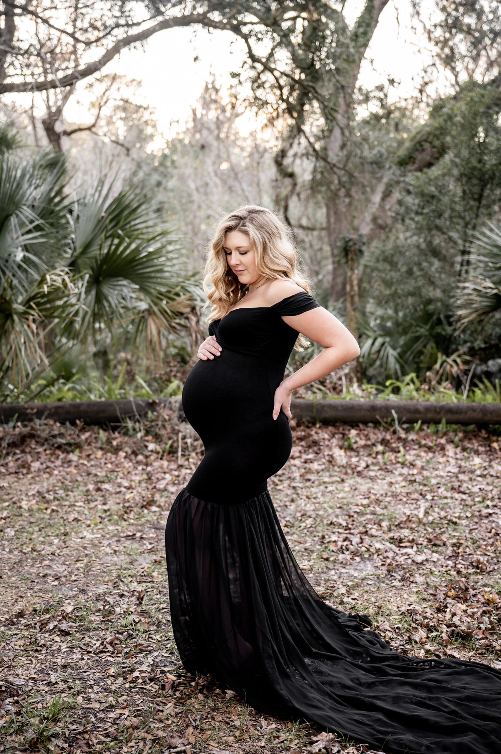 A mother to be in a long black maternity gown stands in a forest gazing down at the bump after using duke prenatal care