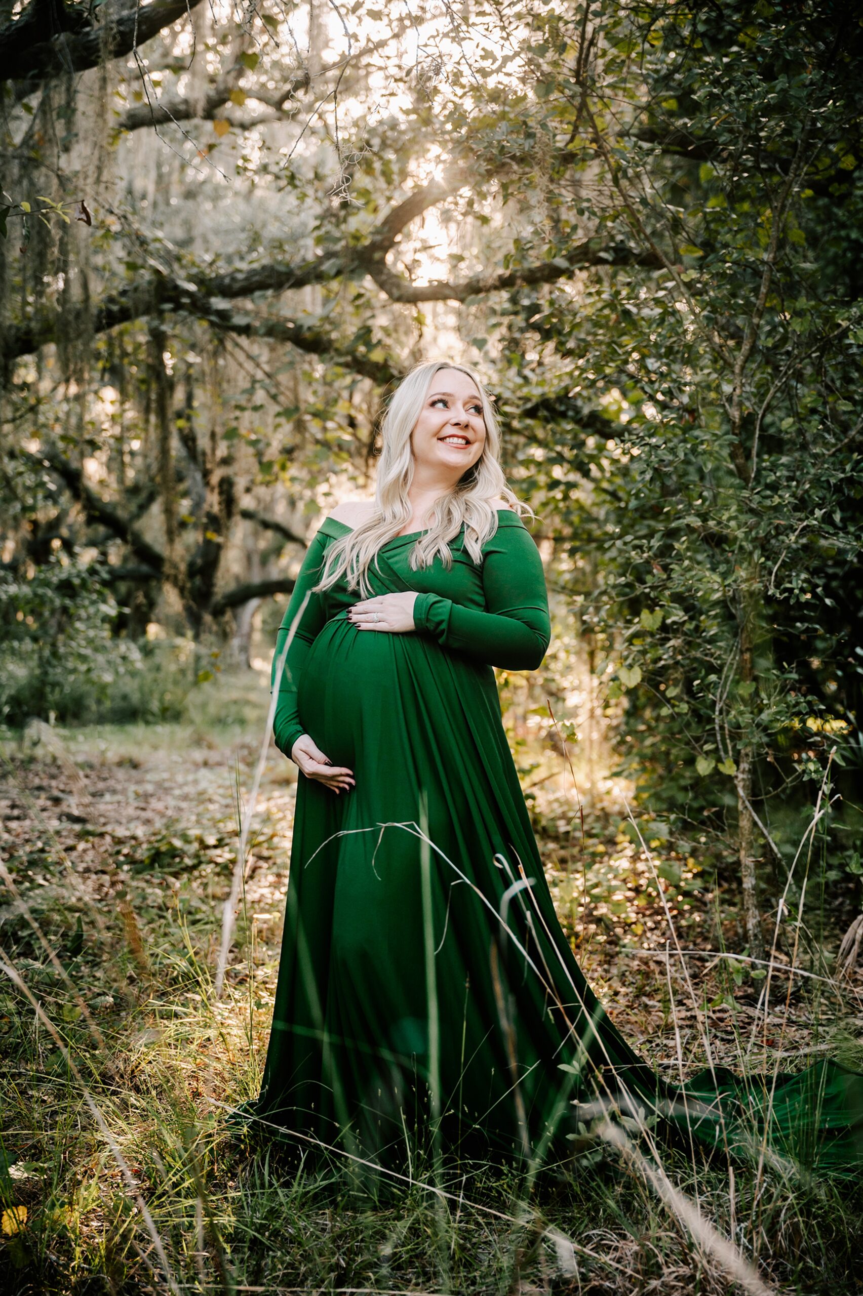 A mom to be stands in a forest at sunset in a green maternity dress while smiling over her shoulder