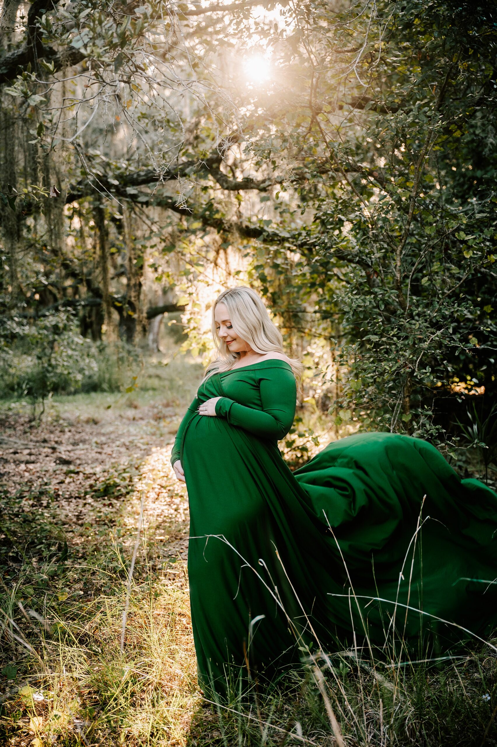 A mother to be in a long flowing green maternity gown smiles down at her bump while standing in a forest at sunset after meeting emerald doulas