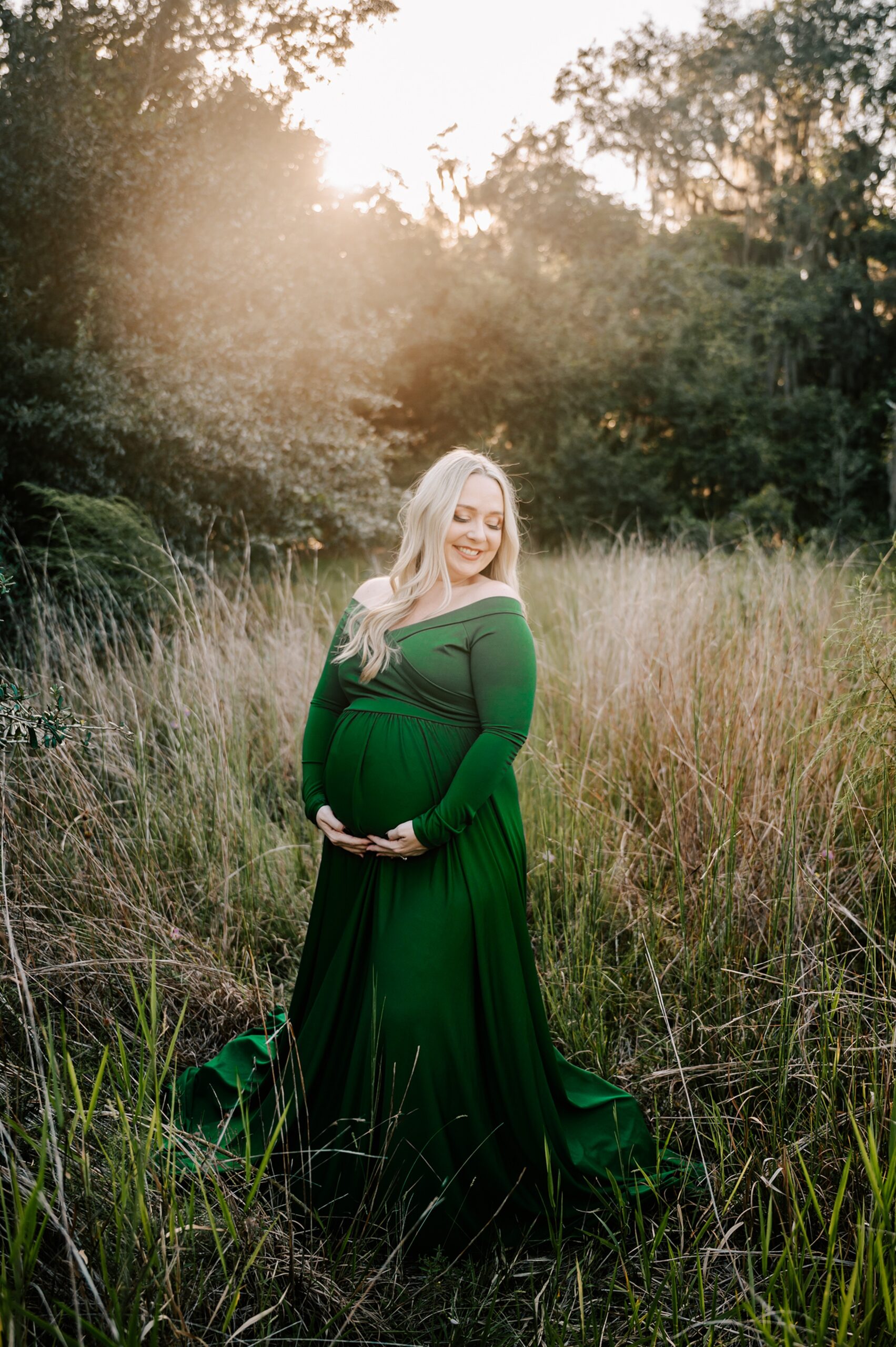 A mother to be in a green maternity gown stands in a field of tall grass at sunset smiling down her shoulder after meeting emerald doulas
