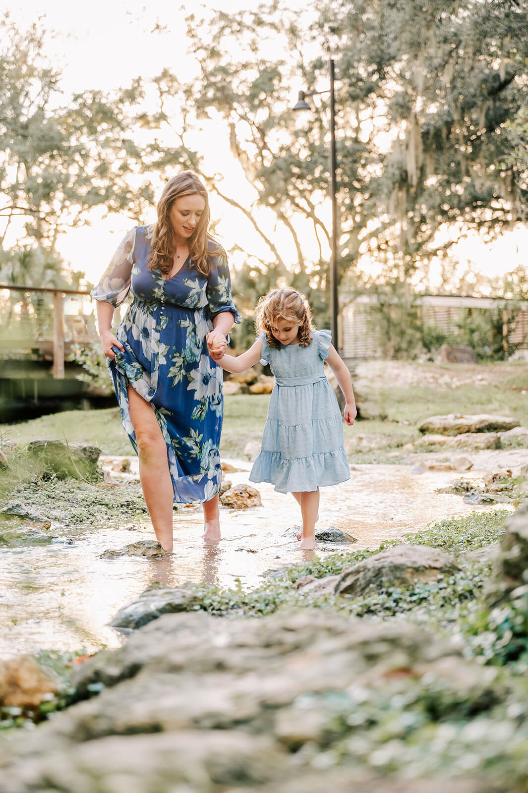 A mother in a blue floral dress walks through a shallow creek holding hands with her daughter in a blue dress at sunset after visiting escape massage in charlotte