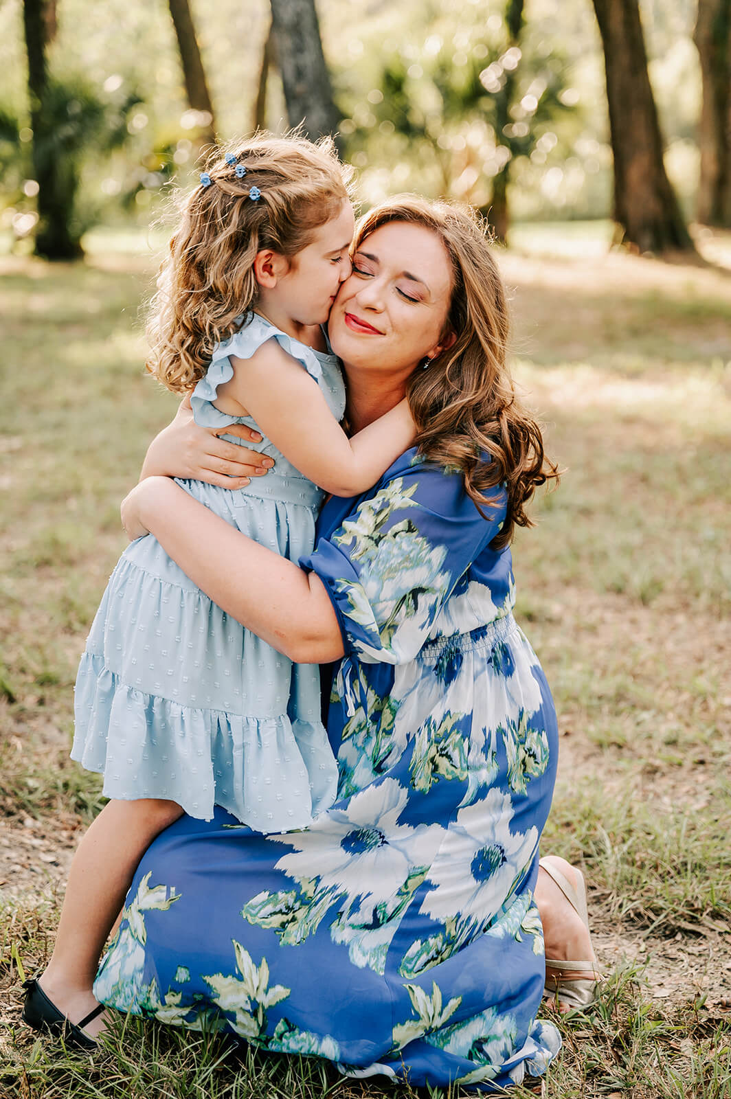 A mother in a blue floral dress hugs her young daughter as she kisses her cheek in a park at sunset after visiting escape massage in charlotte