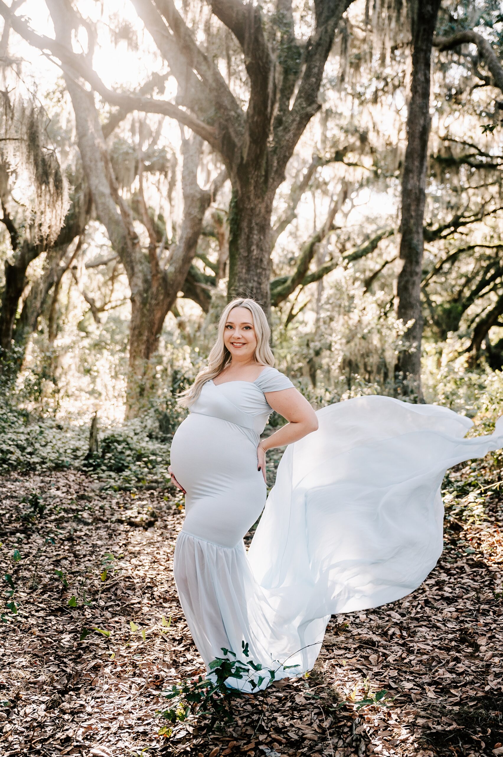 A mother to be stands in a forest with her blue maternity gown train flowing behind her in the wind