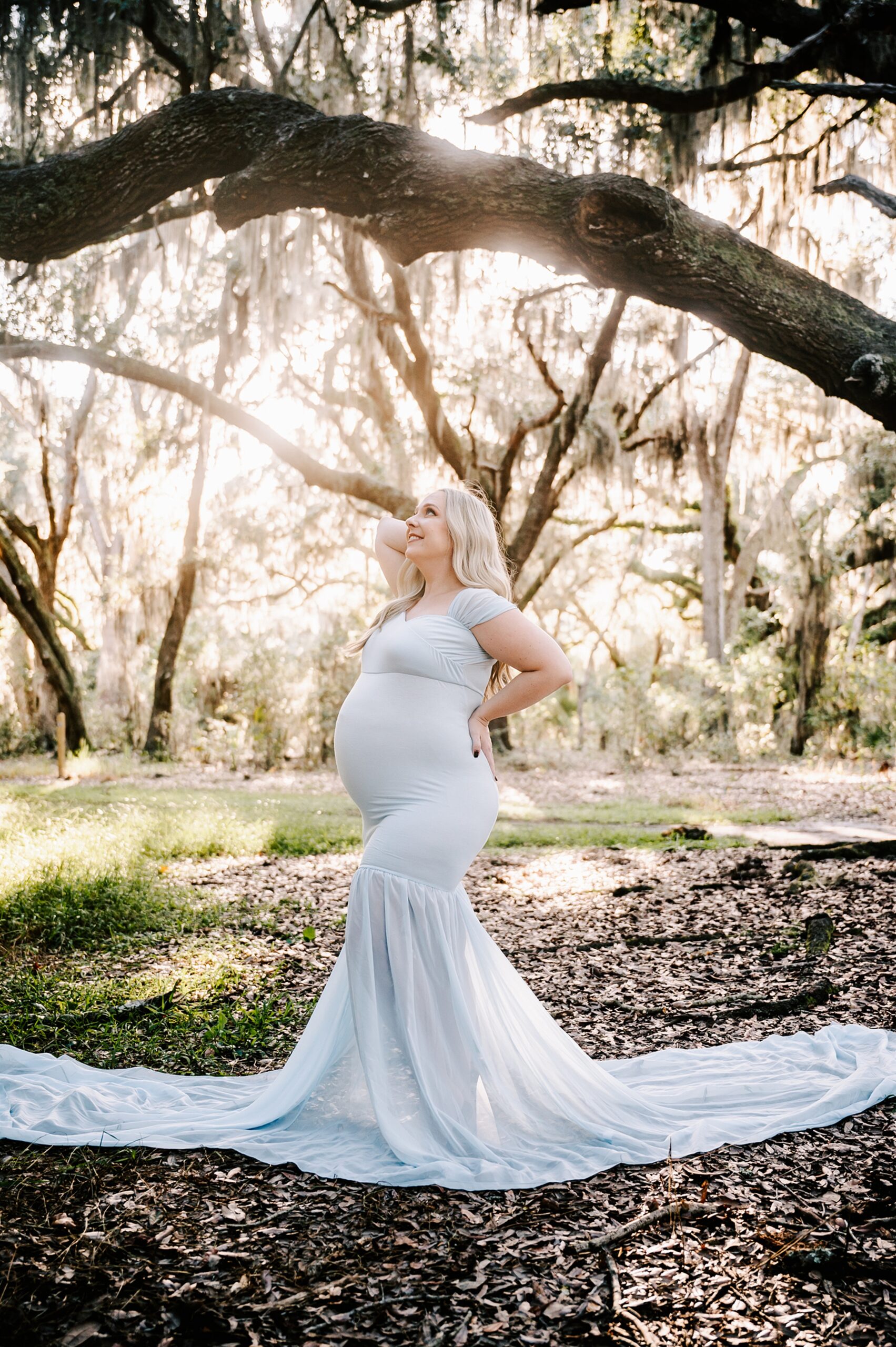 A mother to be stands in a forest at sunset in a blue maternity gown with a hand on her back after visiting forsyth labor and delivery