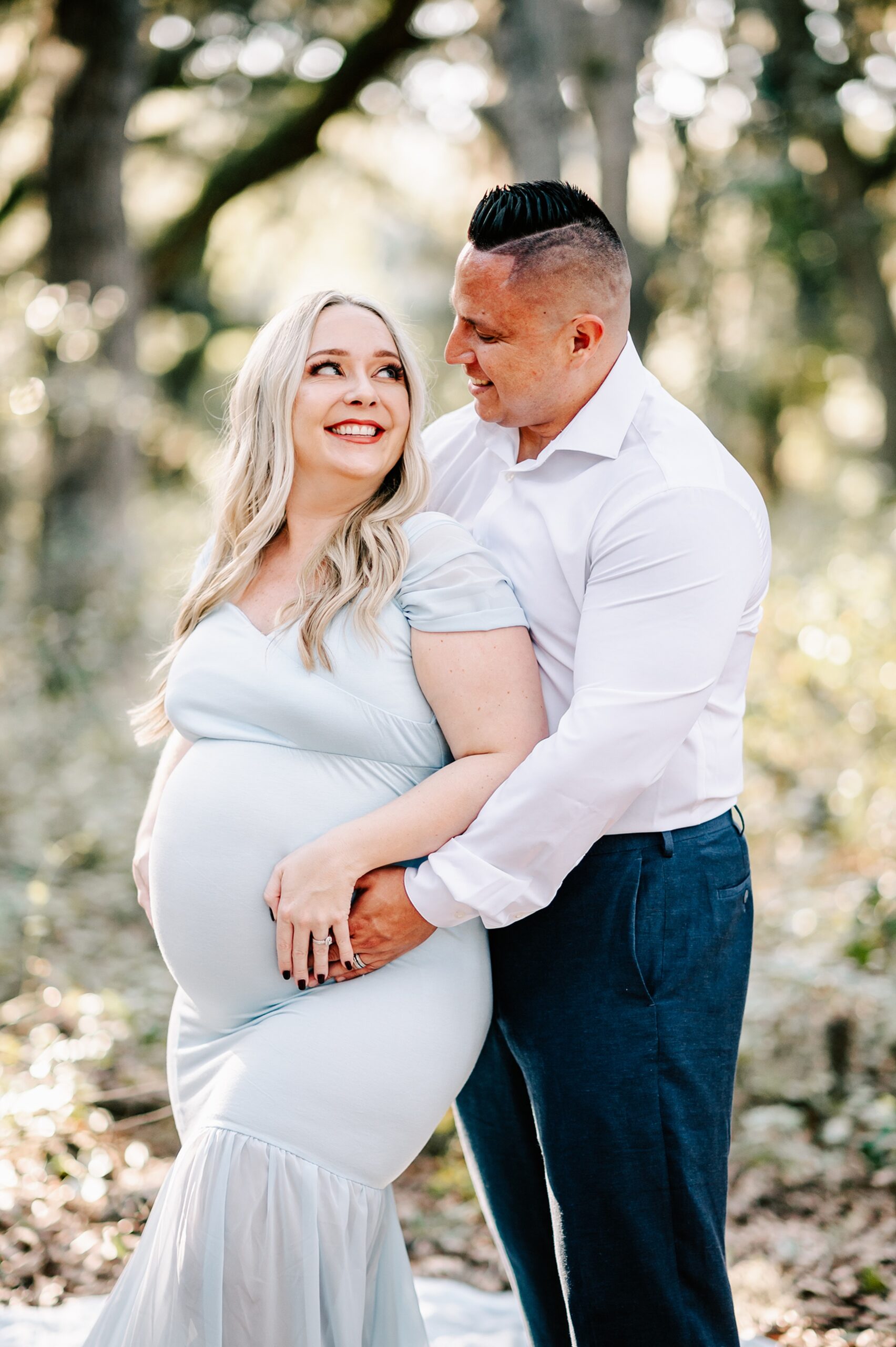 A mother to be Stands in a forest leaning into her husband in a white maternity gown after visiting forsyth labor and delivery