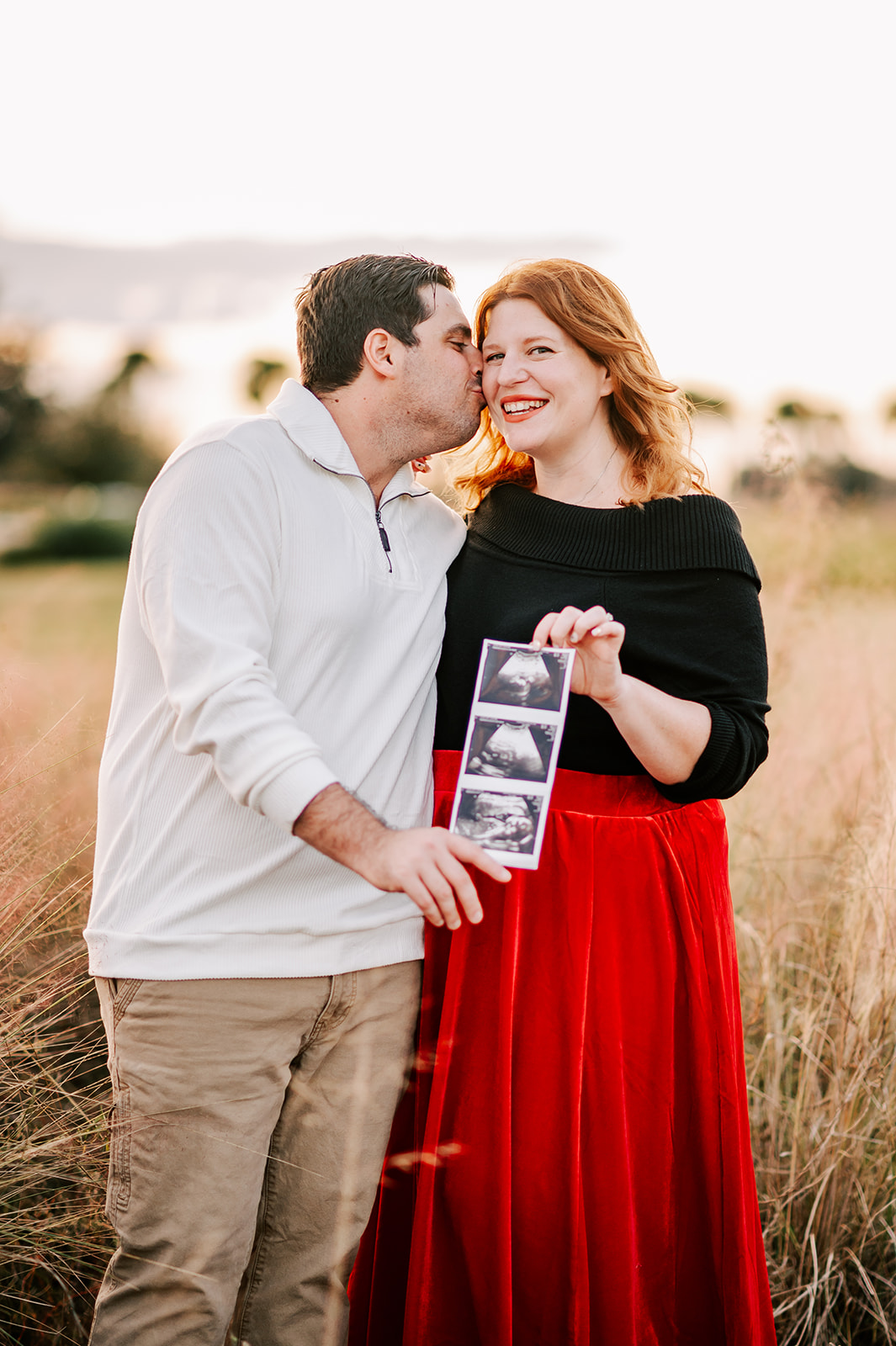 A husband in a white sweater kisses his pregnant wife in a red skirt while standing in a field of tall grass