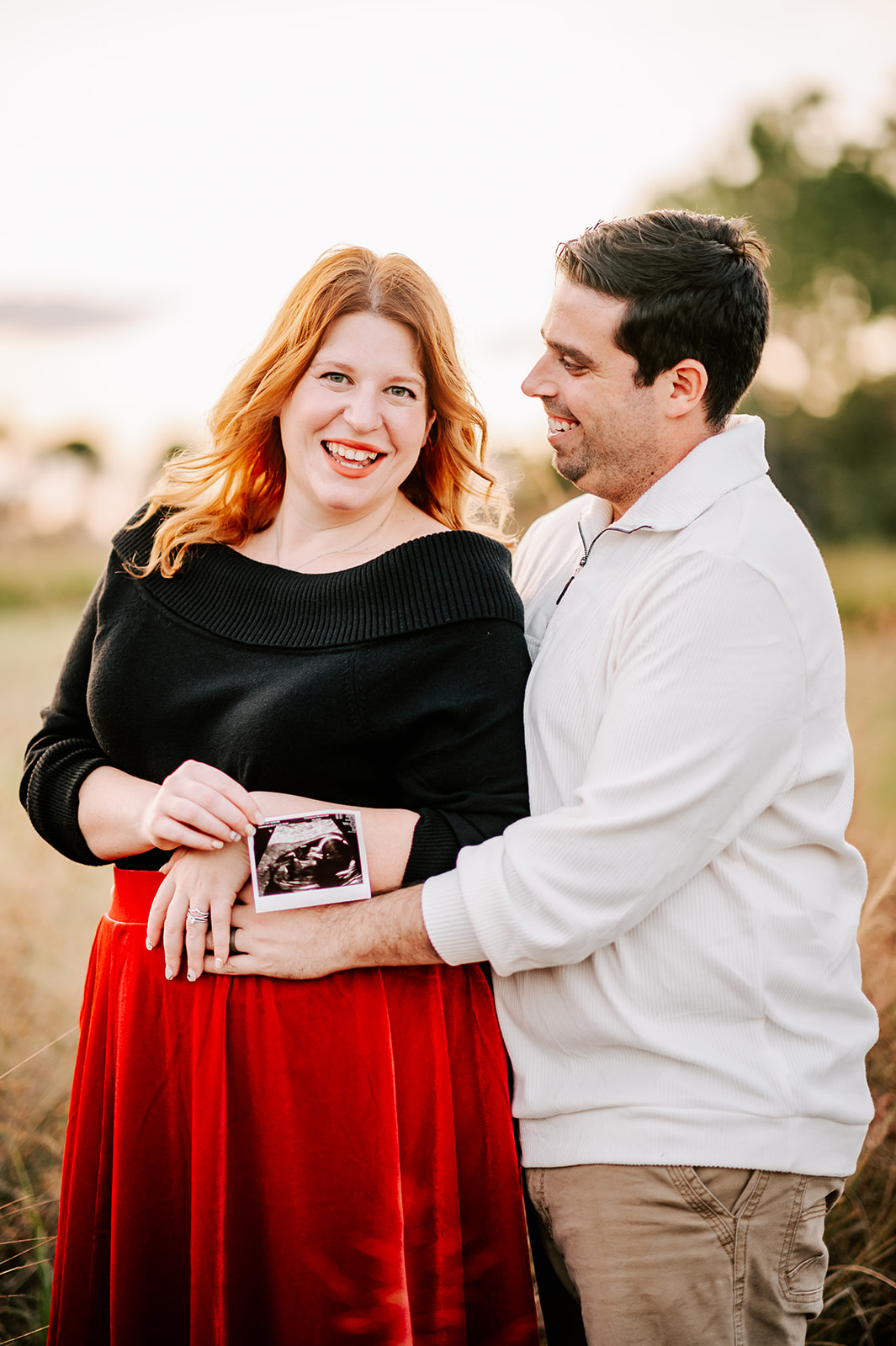 Expecting parents hug while standing in a field at sunset holding a sonogram after meeting Journey of Motherhood