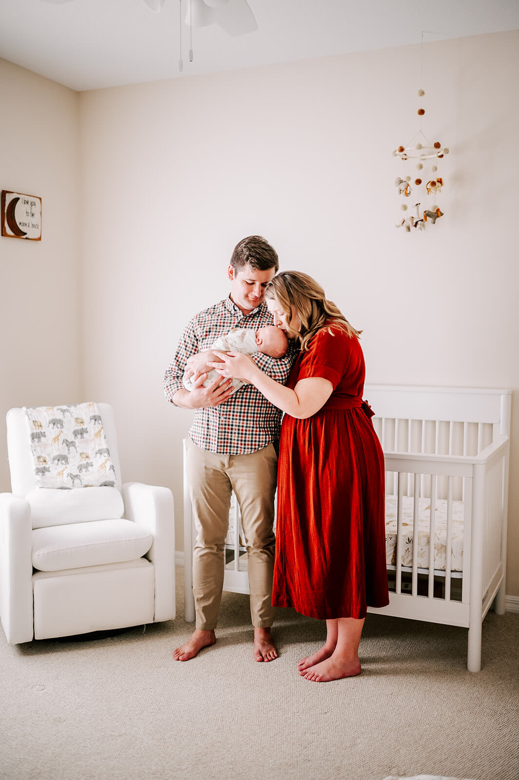 A mother in a red dress kisses her sleeping newborn baby in dad's arms while standing in a nursery