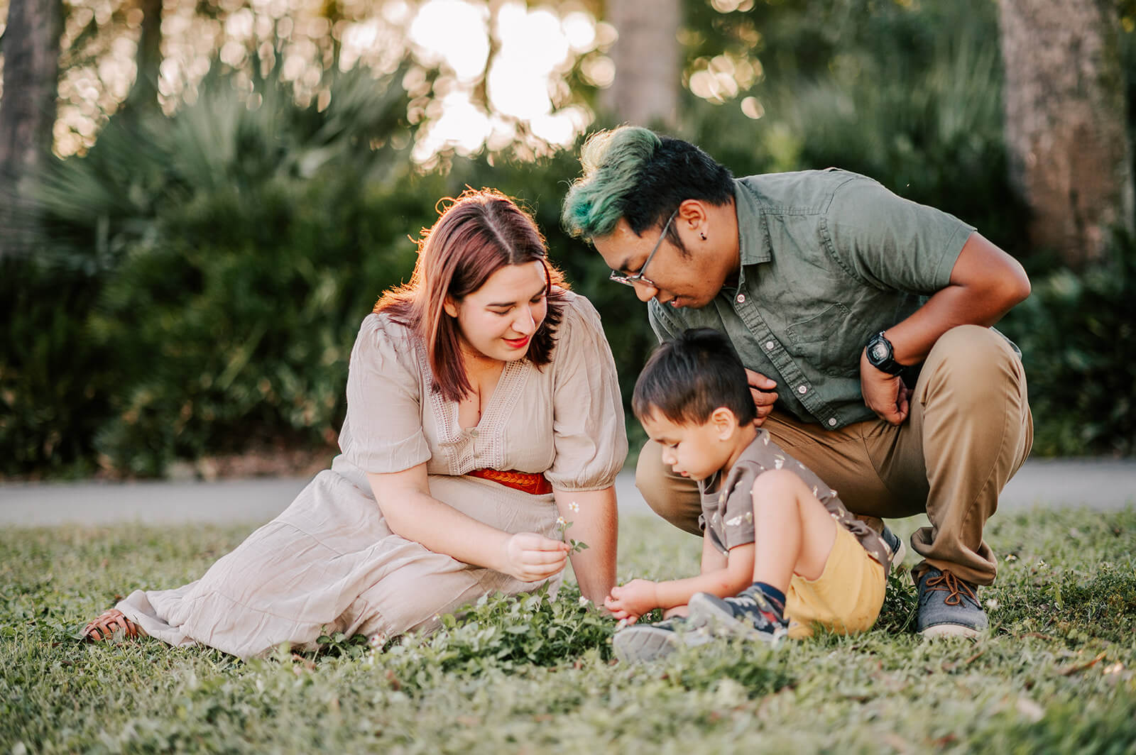 A mom and dad explore some wildflowers in a park lawn with their toddler son
