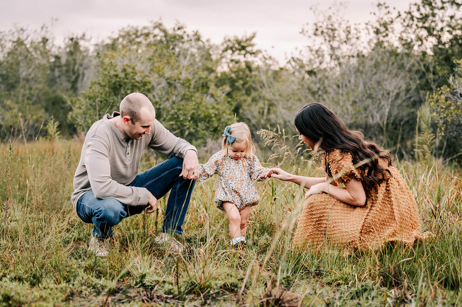 A mom and dad explore a grassy forest field with their toddler daughter