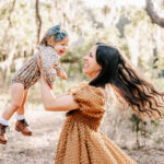 A mother twirls and plays with her toddler daughter in her hands in a park at sunset after visiting massage destination spa